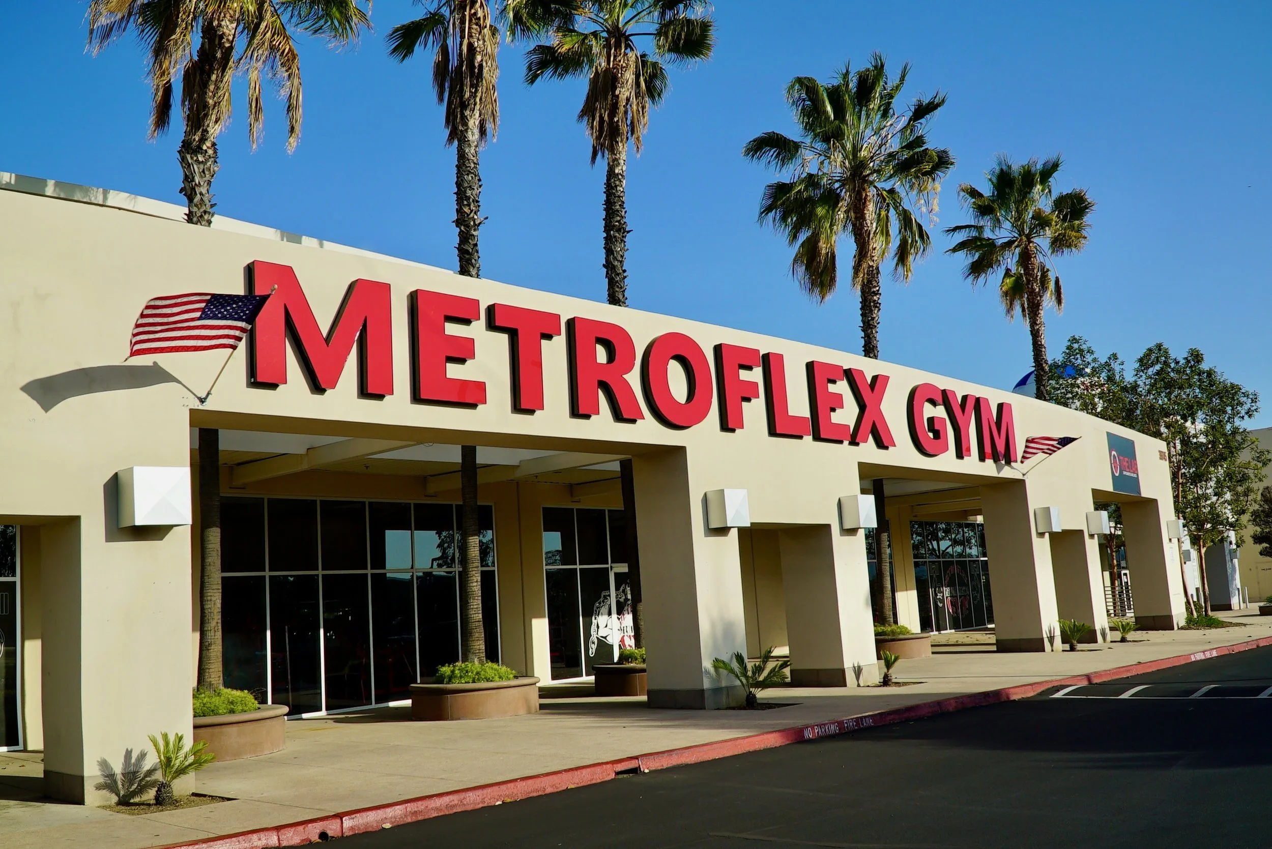 Exterior view of the Metroflex Gym building with a sign in large red letters and American flags, surrounded by palm trees under a clear blue sky.