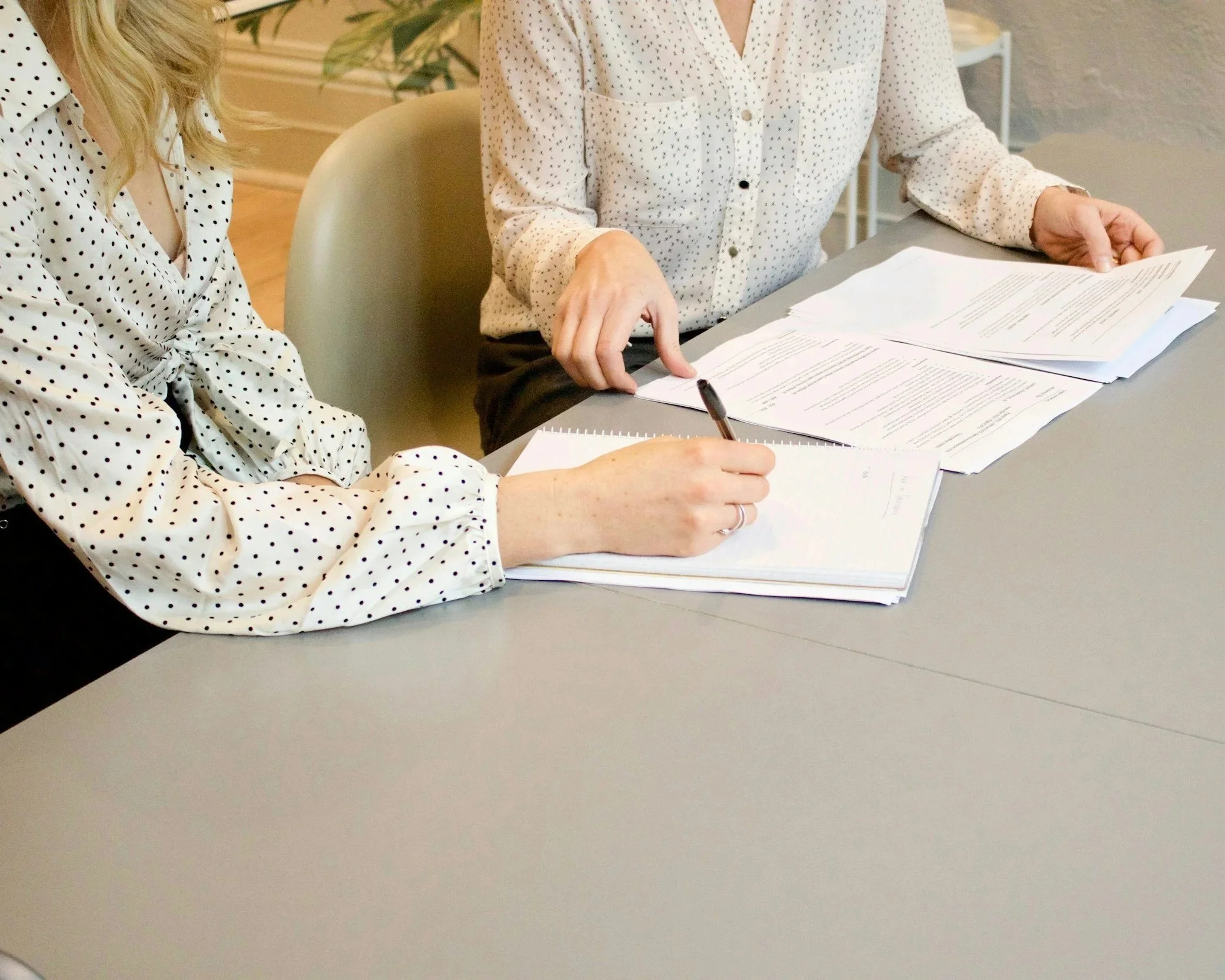 Two women in polka dot blouses sitting at a desk with papers, documents, and a pen, discussing or reviewing paperwork.