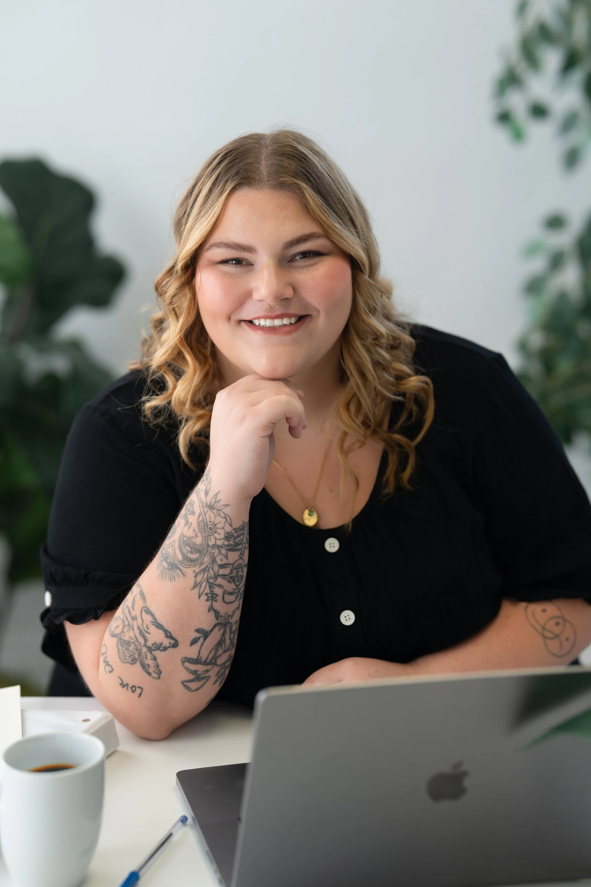 A smiling woman with blonde wavy hair, tattoos on her left arm, wearing a gold necklace with a pendant, sitting at a desk with a laptop, coffee mug, and pen, surrounded by green plants.