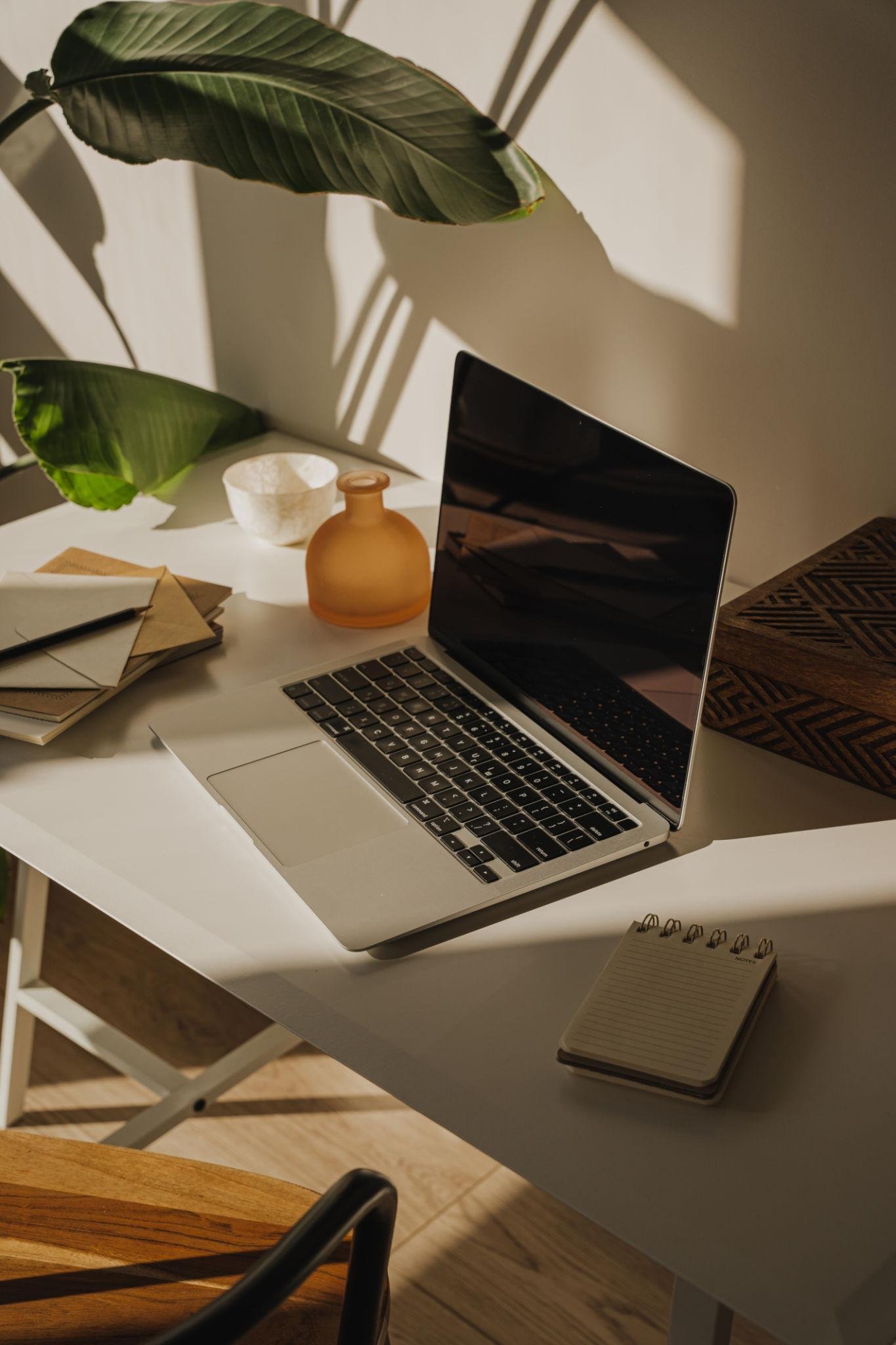 A workspace with a white desk holding a silver laptop, a small notebook, a variety of notebooks, a small white bowl, an orange-brown vase, and large green leaves casting shadows on the wall behind.