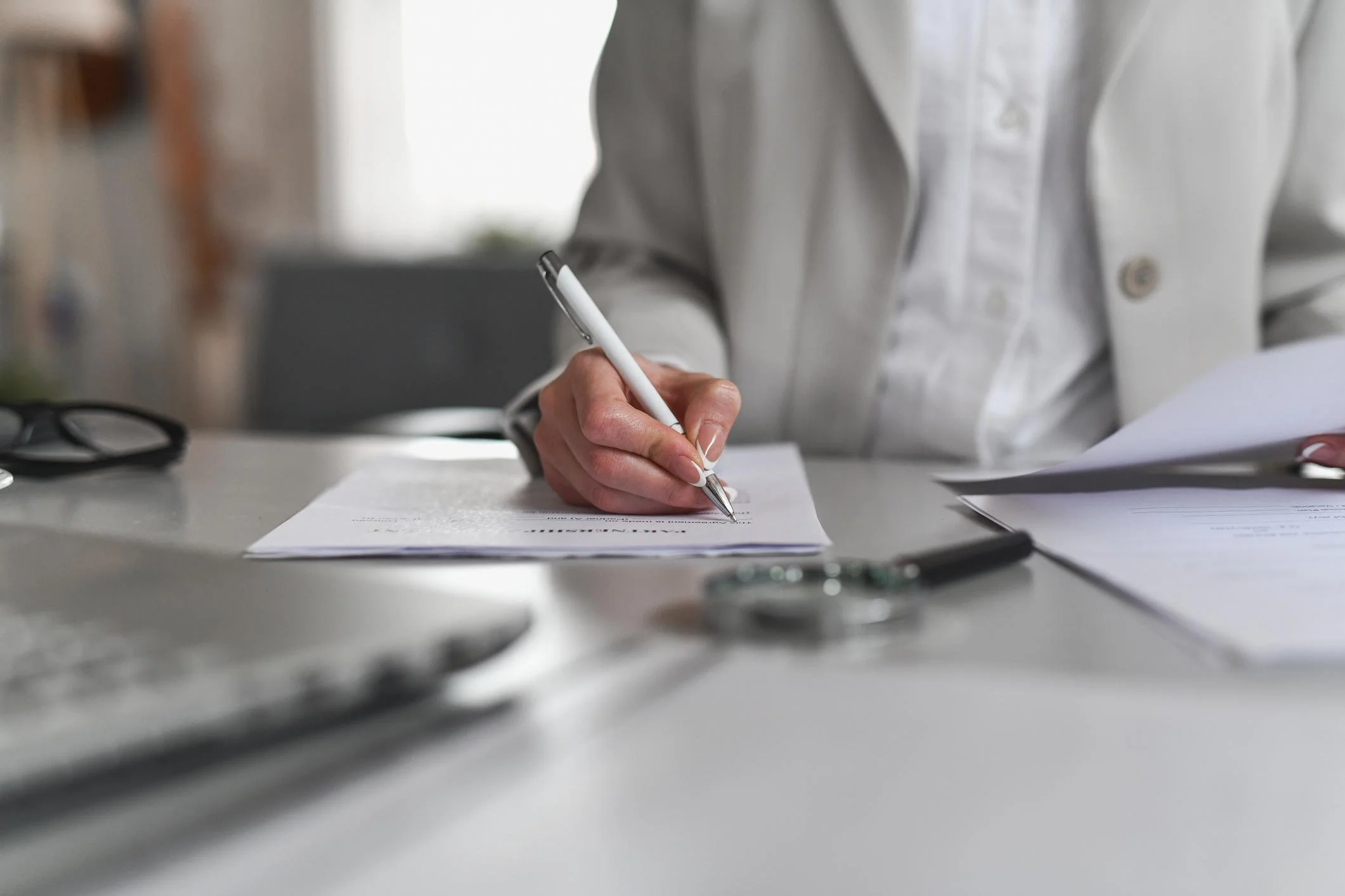 Close-up of a person's hand holding a pen and writing on stacked papers on a desk, with eyeglasses and other papers nearby.