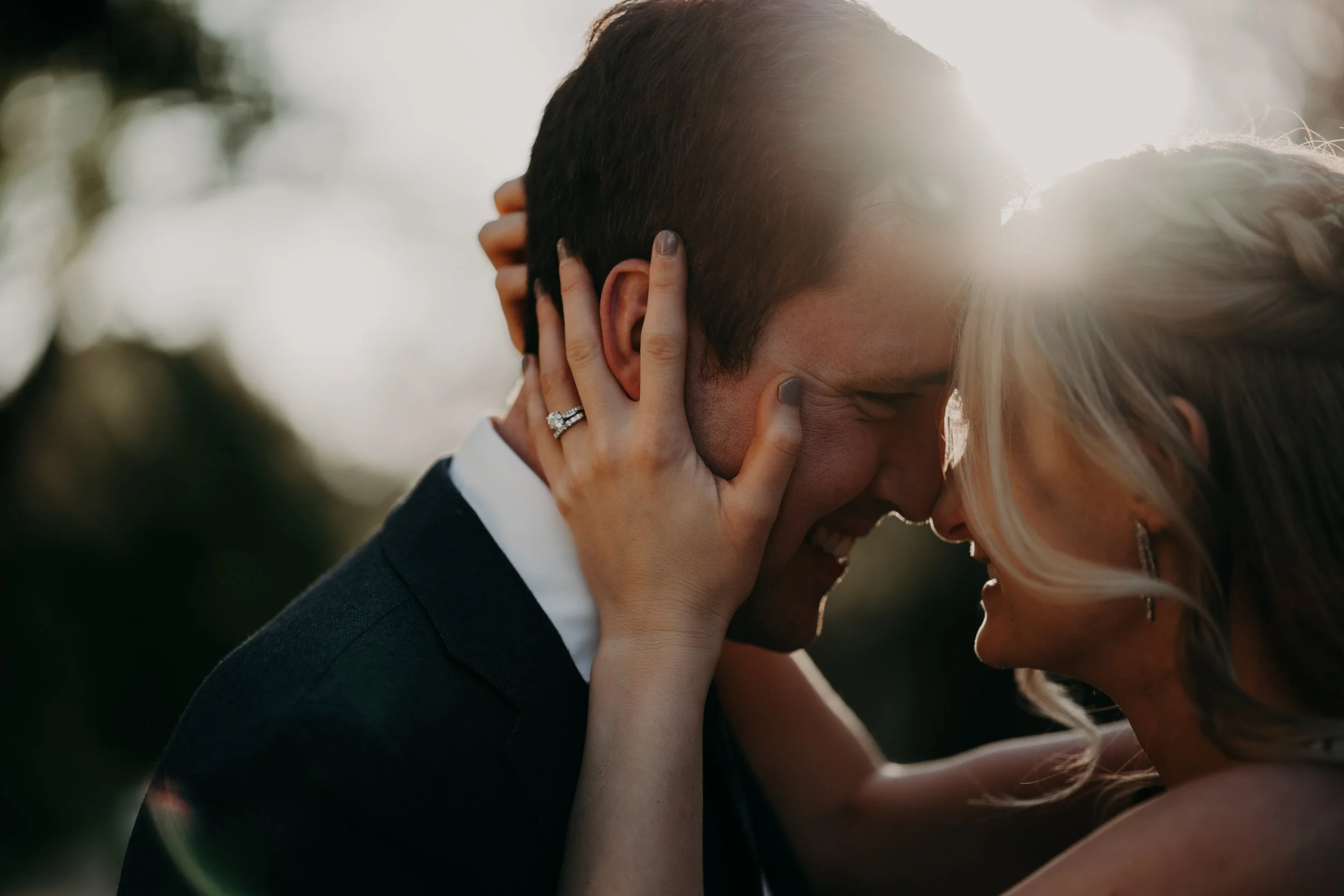 A couple shares a kiss outdoors with sunlight shining behind them.