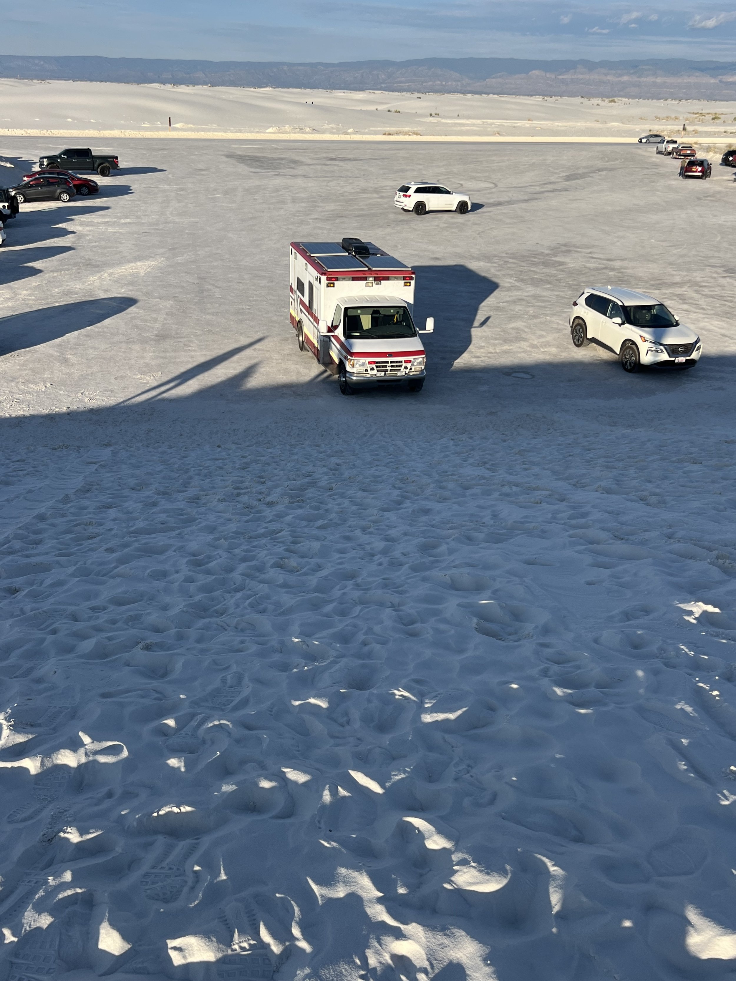 A wide view of a snowy landscape with cars and an emergency vehicle parked on the snow, some mountains in the distance under a partly cloudy sky.