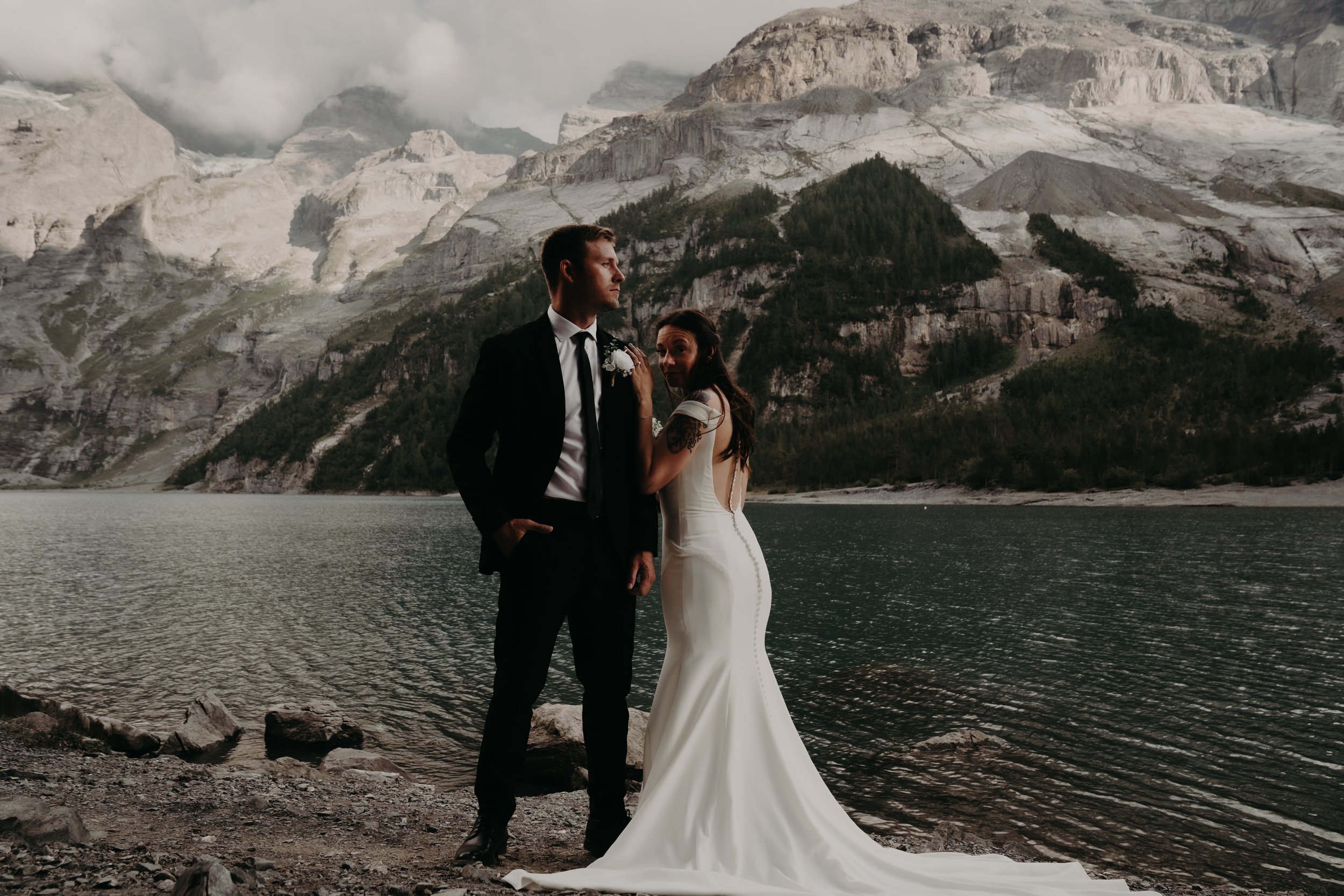 A bride and groom stand by a lake with mountainous terrain in the background. The groom wears a black tuxedo with a white shirt and black tie. The bride wears a white wedding gown and has tattoos on her left arm. They are standing close together, with the bride smiling and the groom looking to the side.