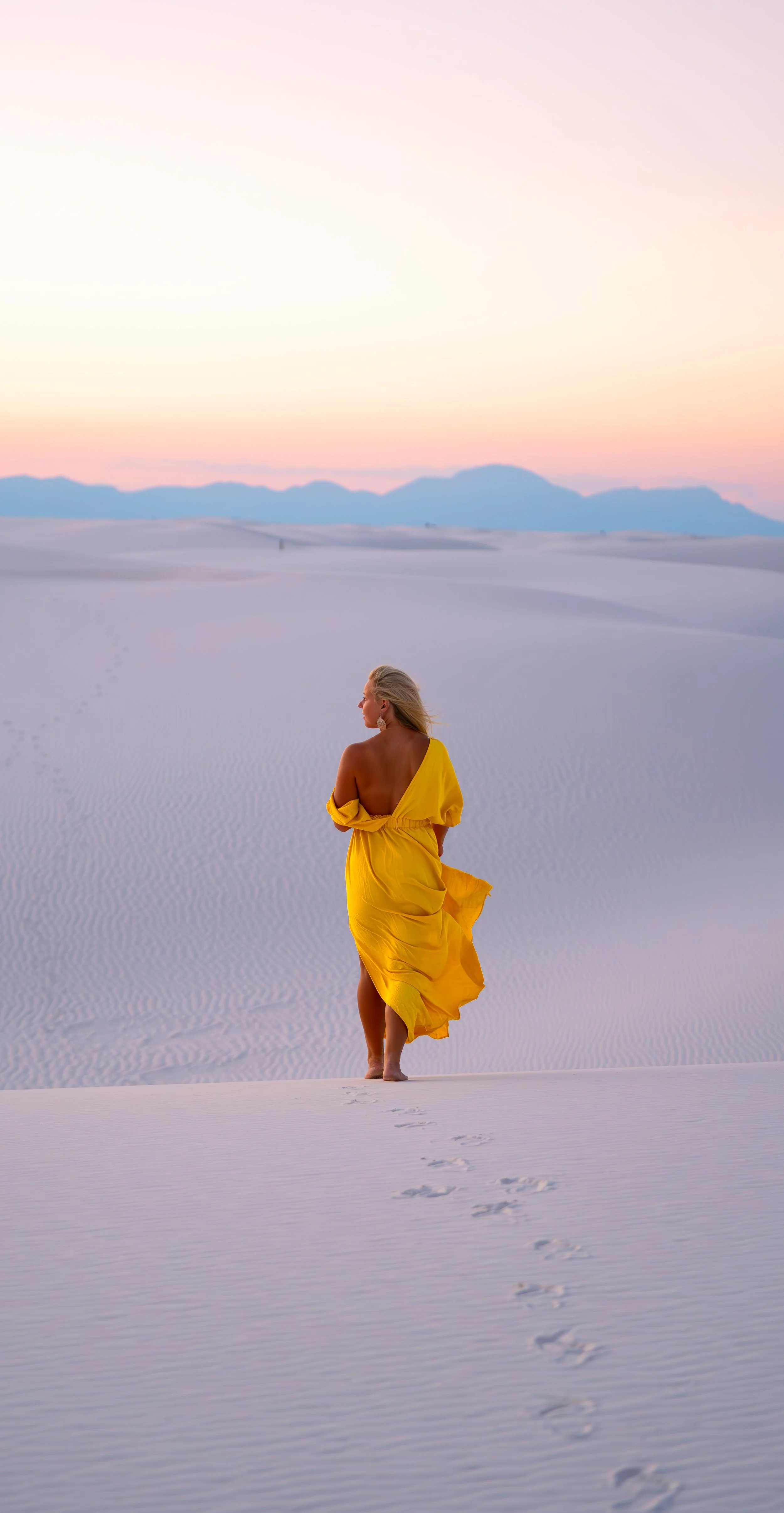A woman in a yellow dress walking barefoot across white sand dunes at sunset, with mountains in the distance.