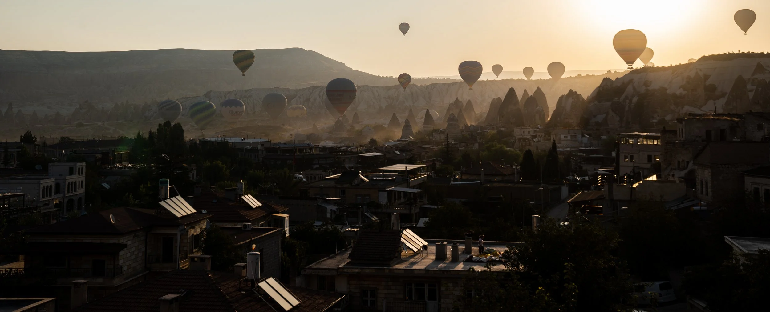 Hot air balloons floating over a town at sunrise with mountainous terrain in the background.