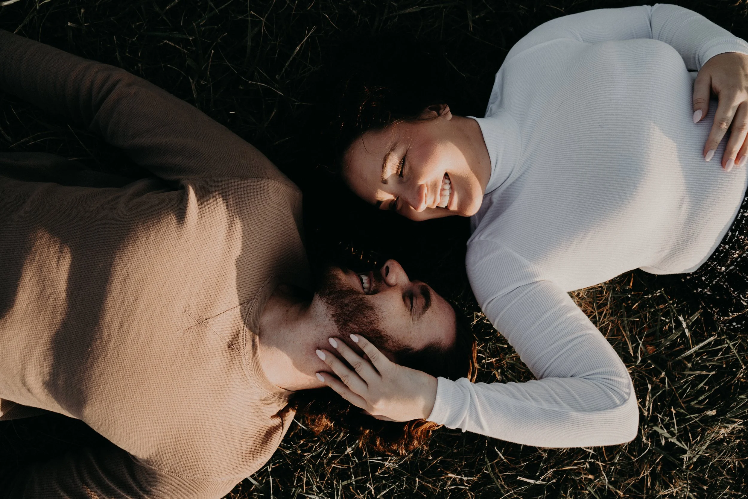 A woman and a man lying on the ground outdoors, smiling and looking at each other. The woman is wearing a white long-sleeve shirt, and the man is wearing a tan shirt. They appear happy and relaxed.