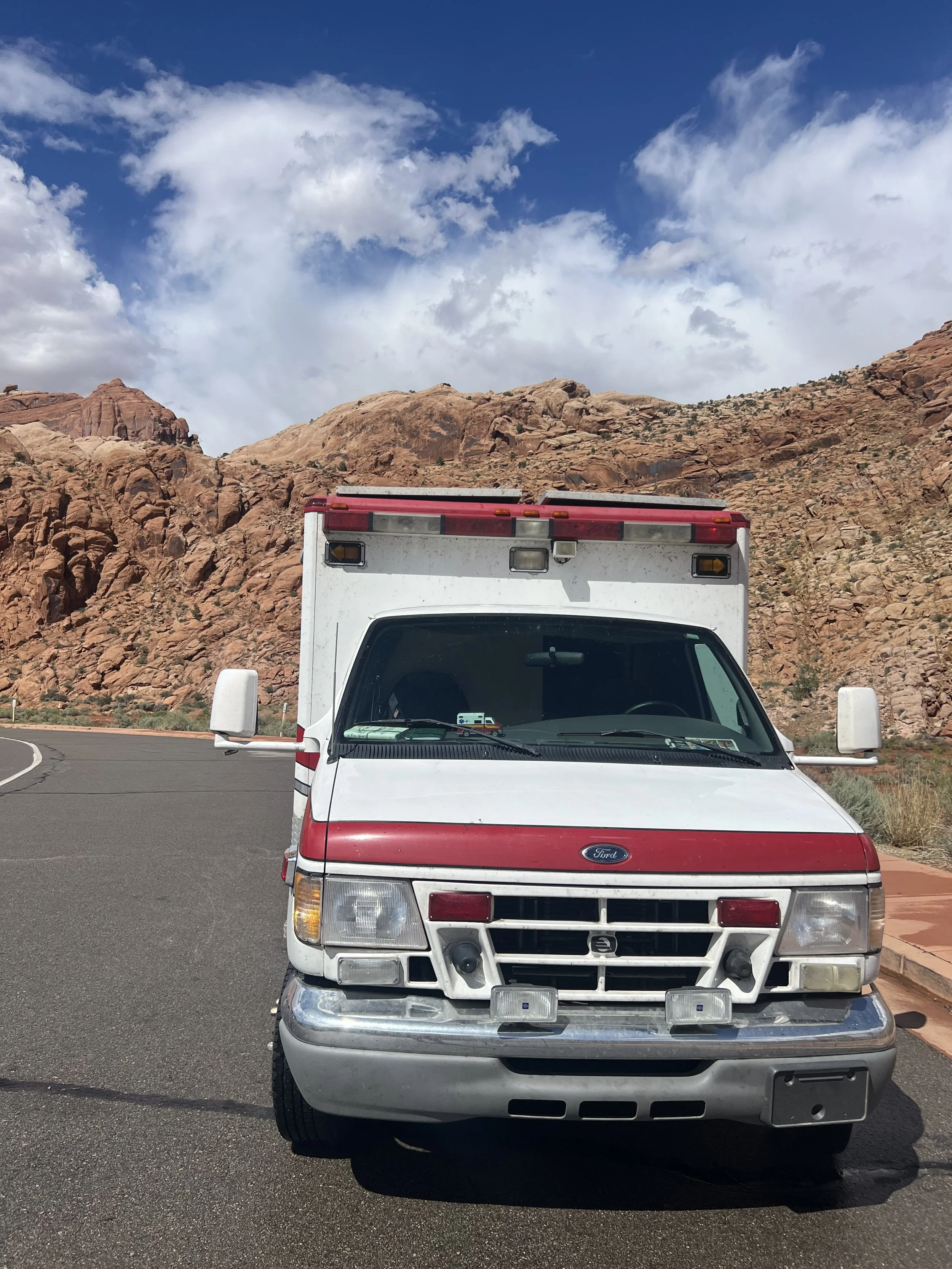 Front view of a white vintage Ford van with red accents parked on a paved road, with a rocky desert mountain landscape and a blue sky with scattered clouds in the background.  Adventure elopement.  converted ambulance.  van life.