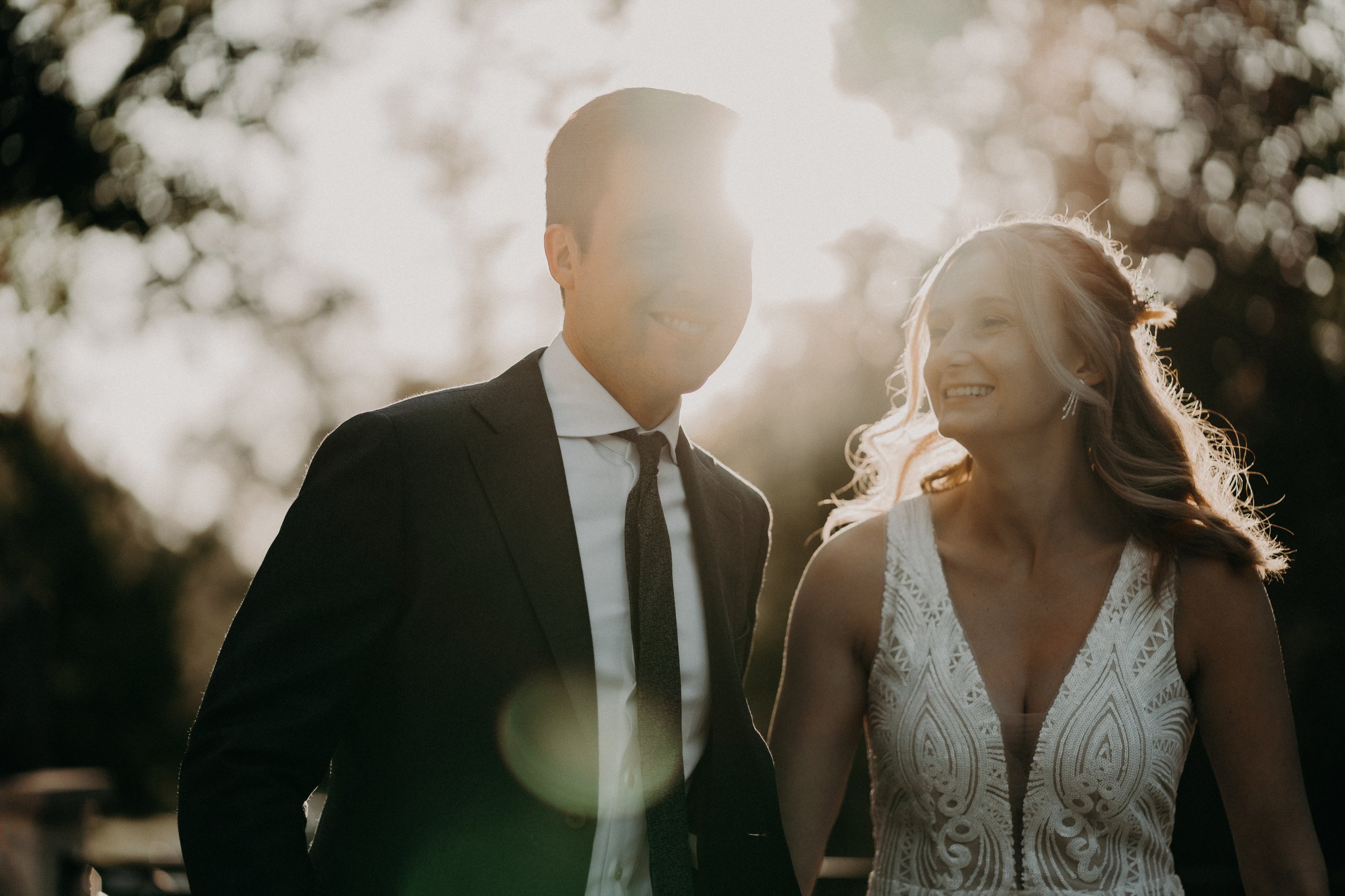 A smiling man in a suit and a woman in a white lace dress, both outdoors with sunlight filtering through trees in the background.