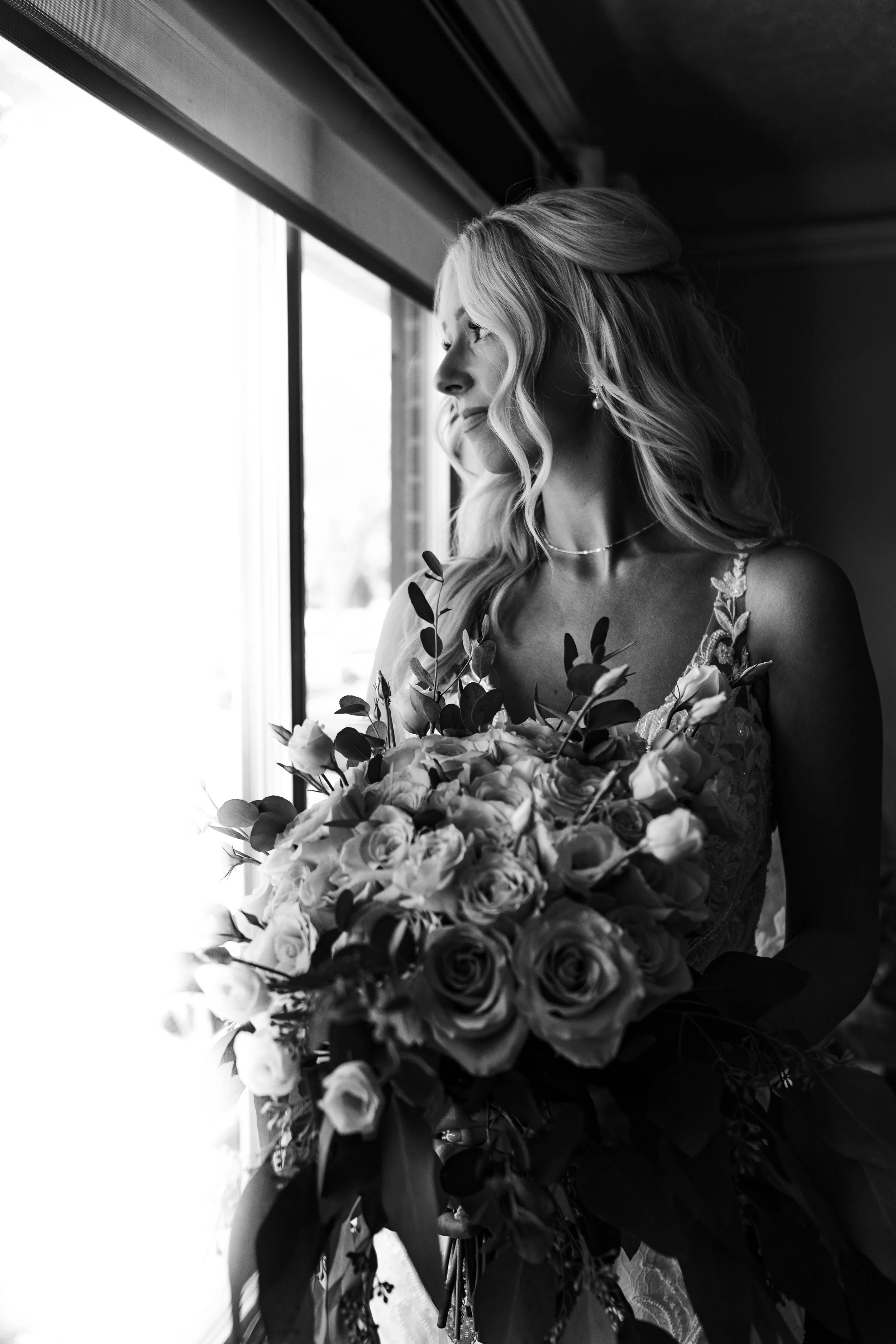 A woman with wavy blonde hair gazes out of a window, holding a large bouquet of flowers, dressed in a lace dress, with pearl earrings and necklace.