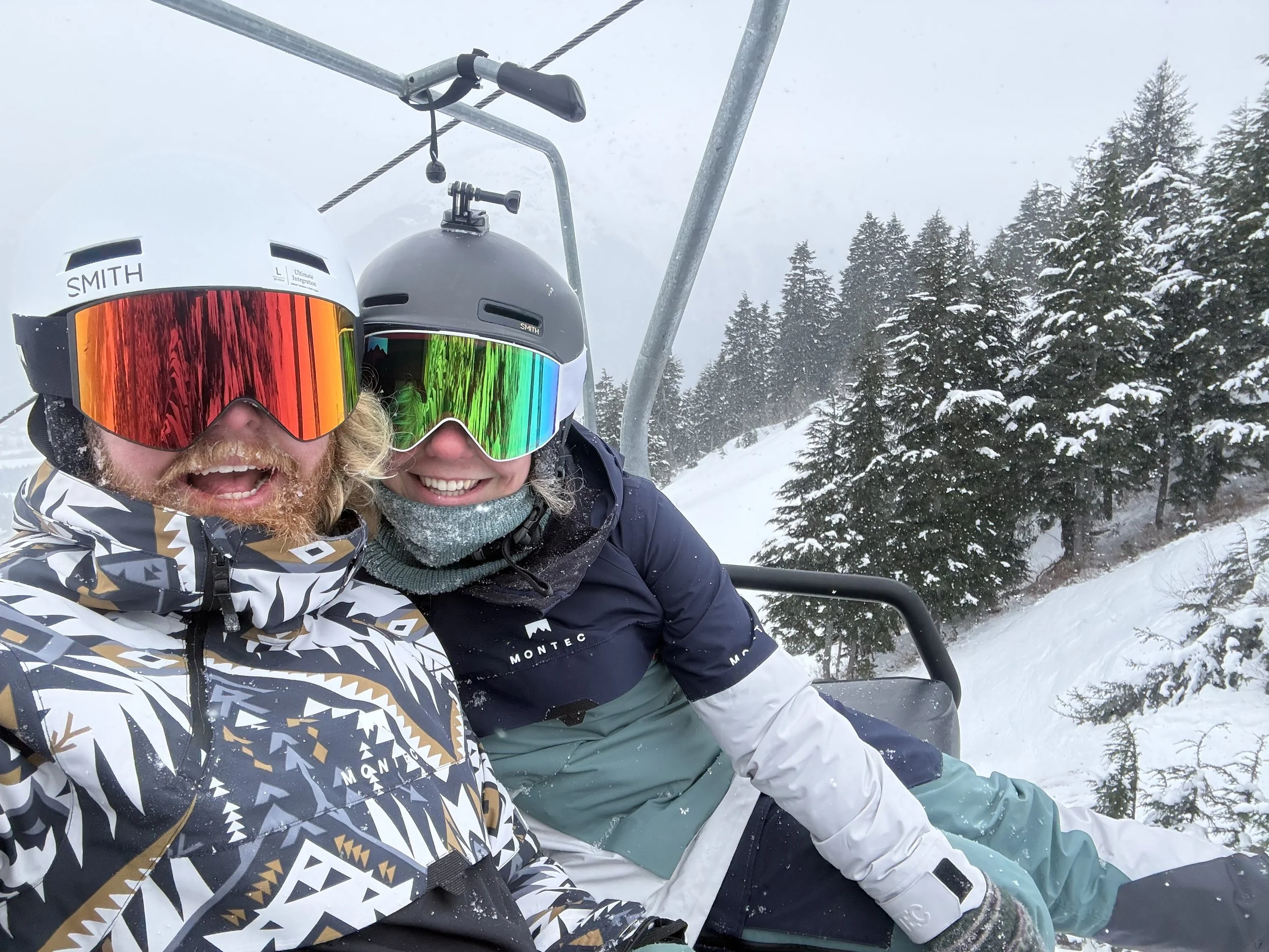 Two skiers wearing helmets and goggles taking a selfie on a snowy mountain with pine trees in the background.