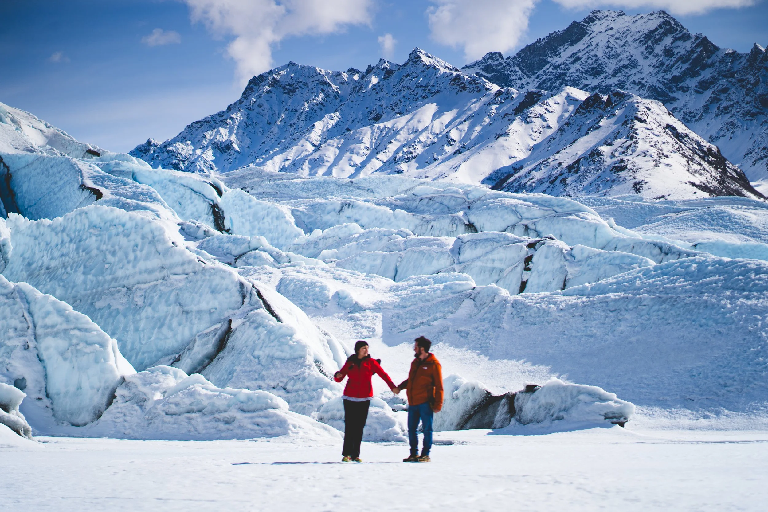 Two people holding hands and talking in a snowy landscape with large ice formations and snow-covered mountains in the background.