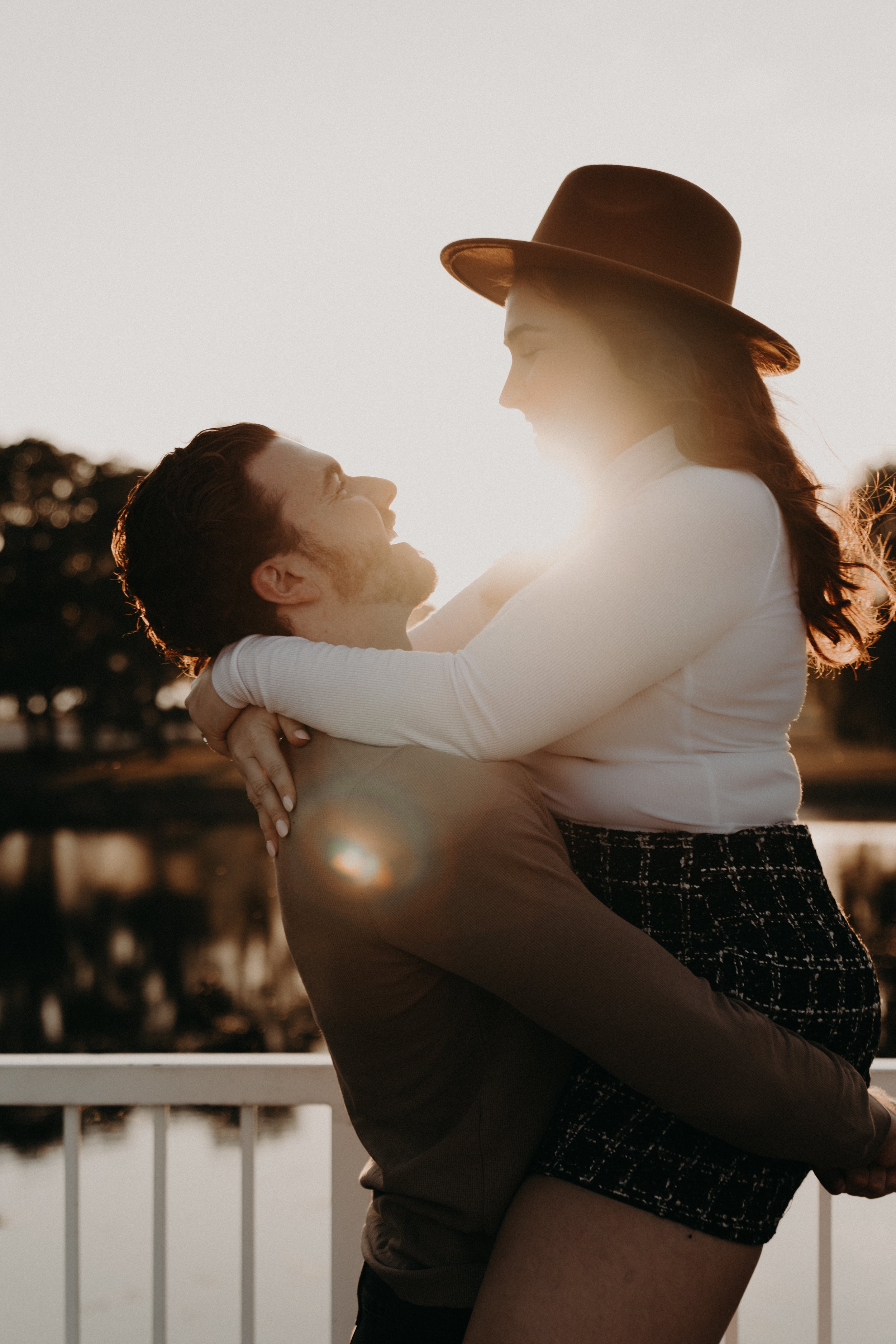 A man and woman hugging outdoors at sunset, with the woman wearing a wide-brimmed hat and the city skyline blurred in the background.