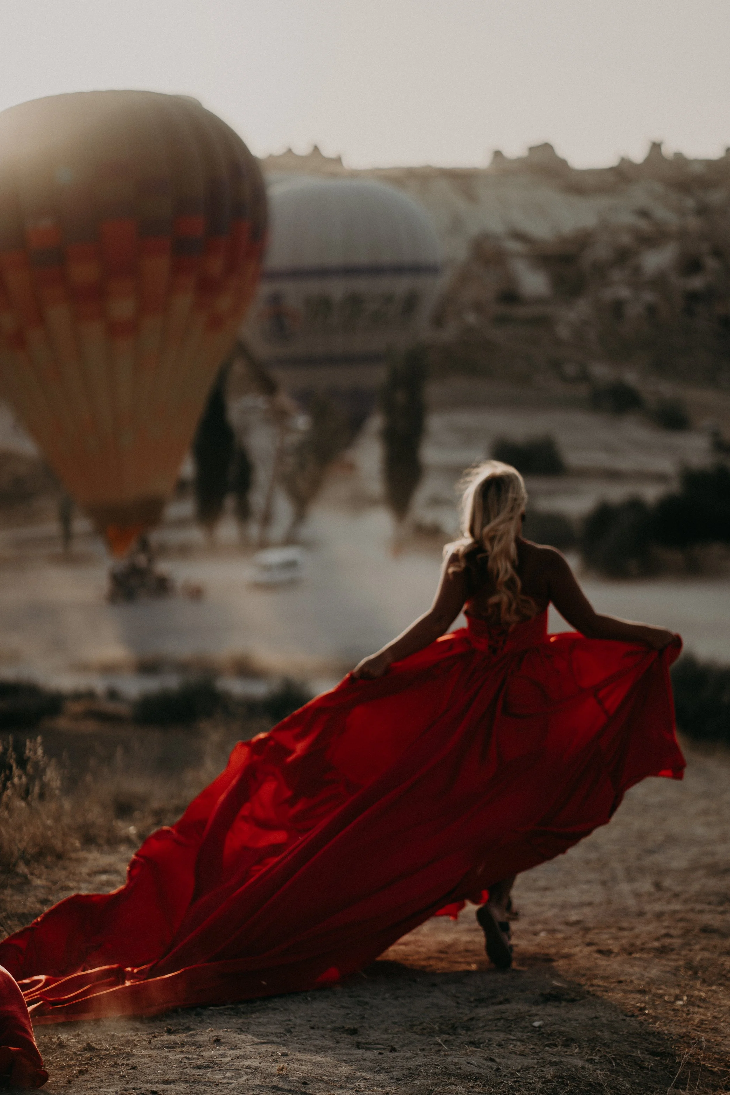 A woman with long blonde hair sitting cross-legged on the ground wearing a flowing red dress, with hot air balloons in the background at sunset.