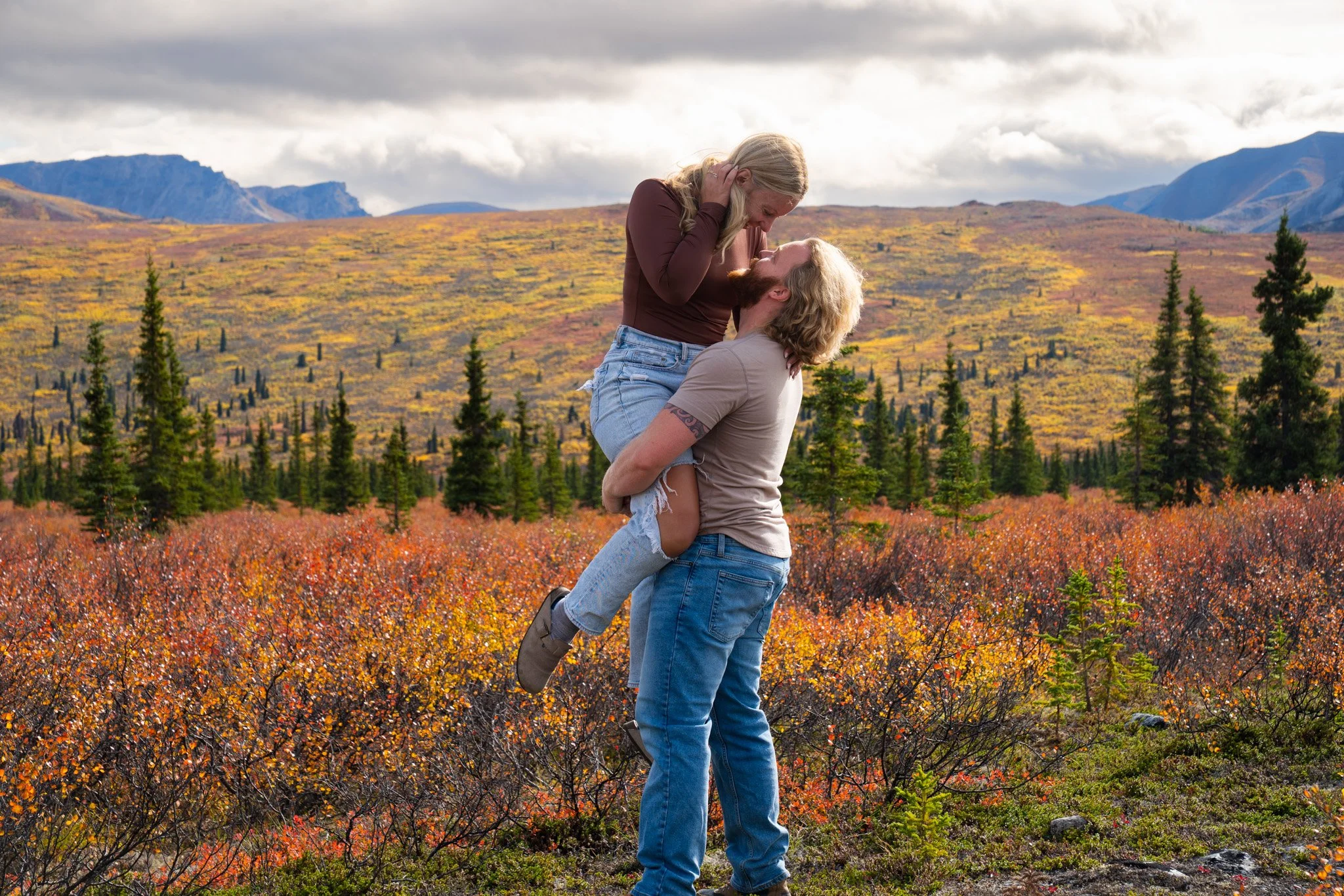 A couple embracing in a field with autumn-colored foliage, mountains in the background, and cloudy skies.