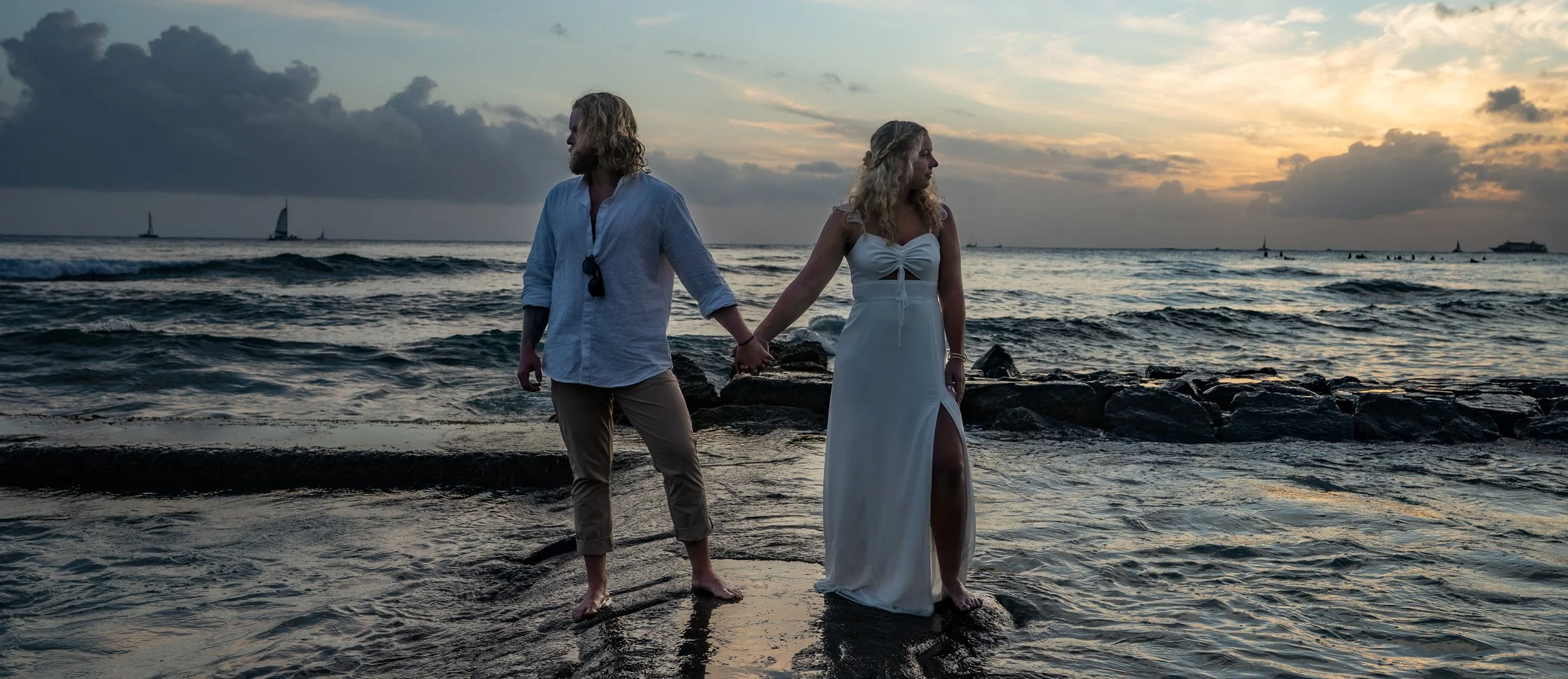A couple holding hands on the beach at sunset, with sailboats in the distance.