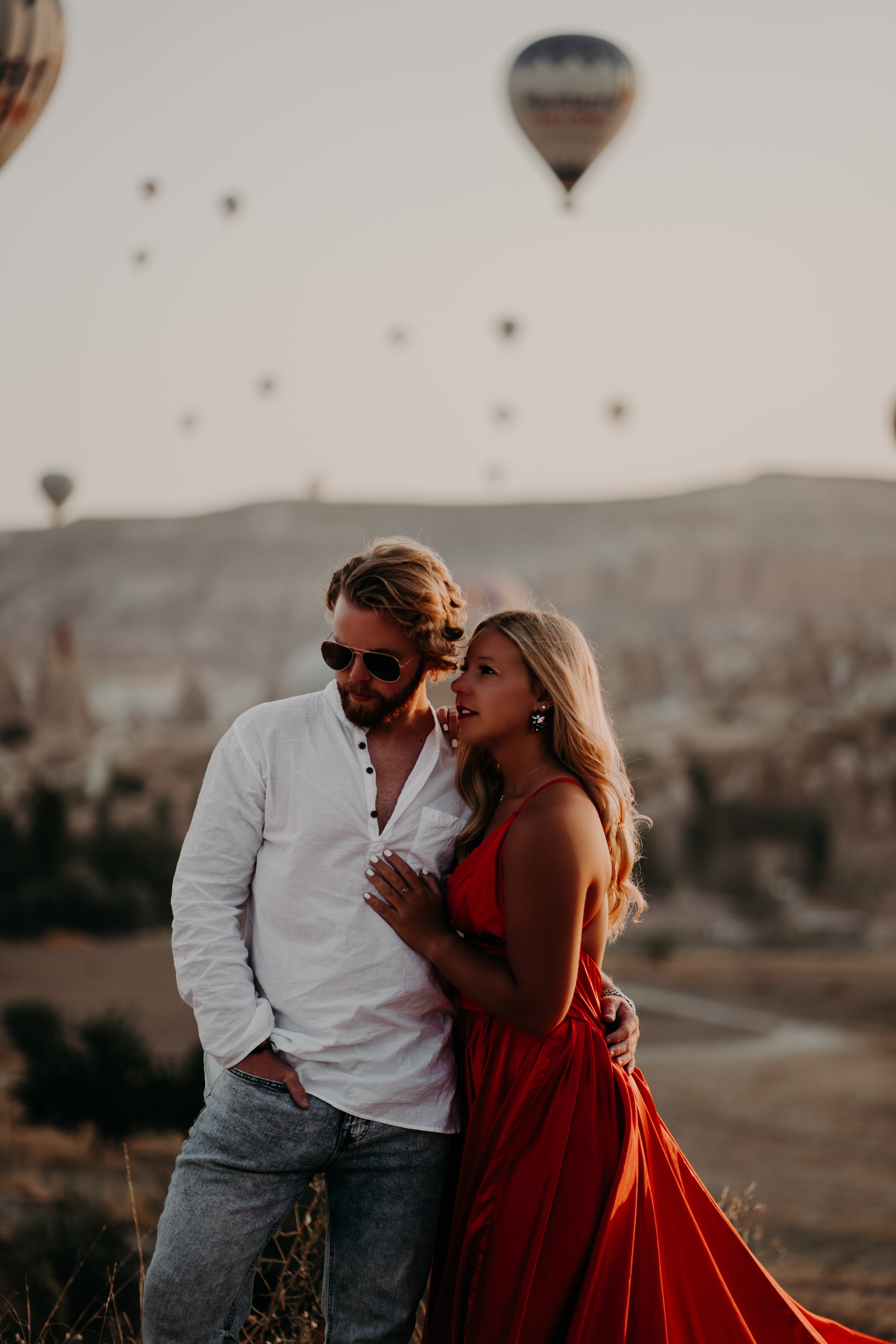 A man and woman standing close together outdoors with hot air balloons in the sky behind them during sunset.