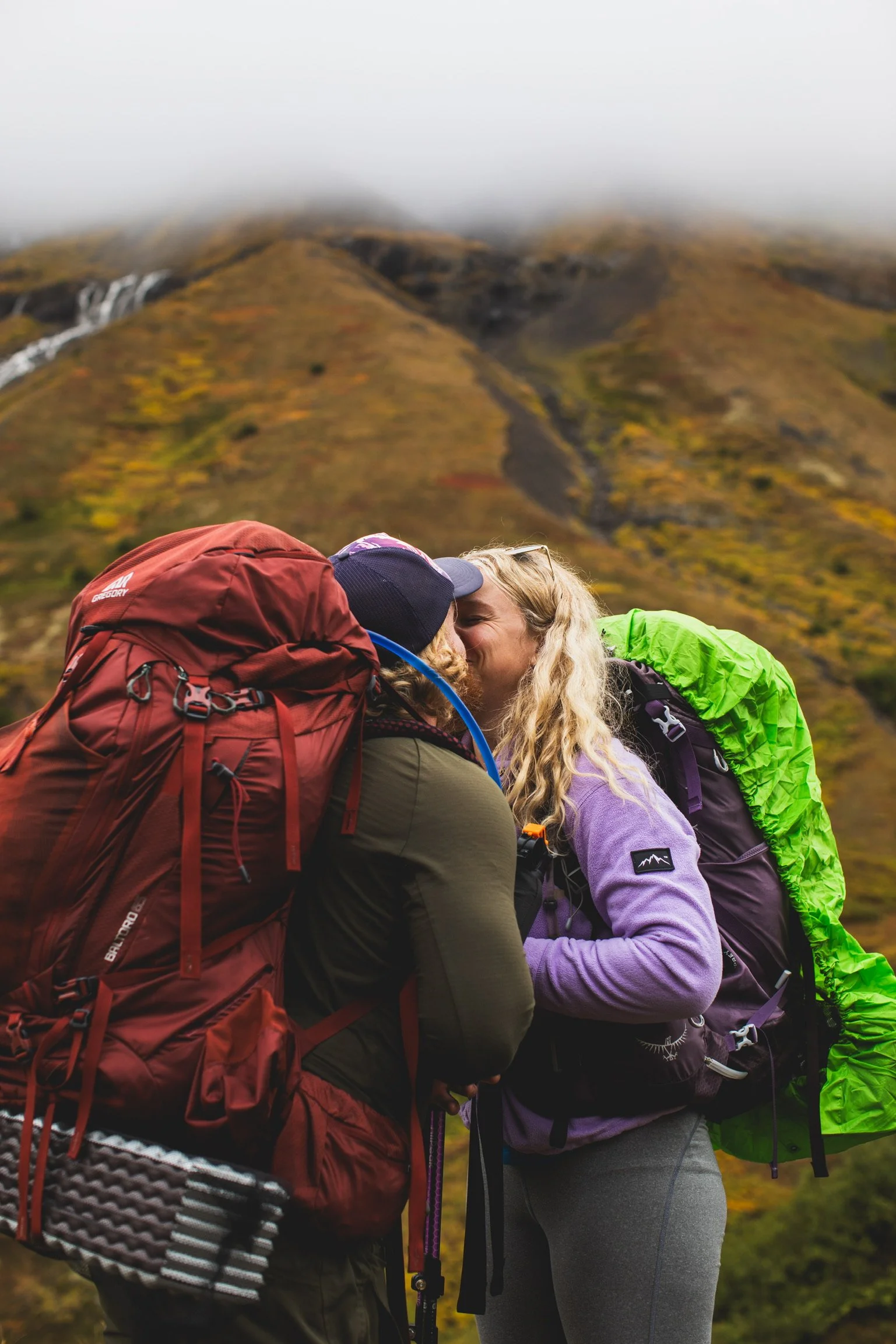 An engaged couple sharing a kiss outdoors during a hike with mountains and cloudy sky in the background.