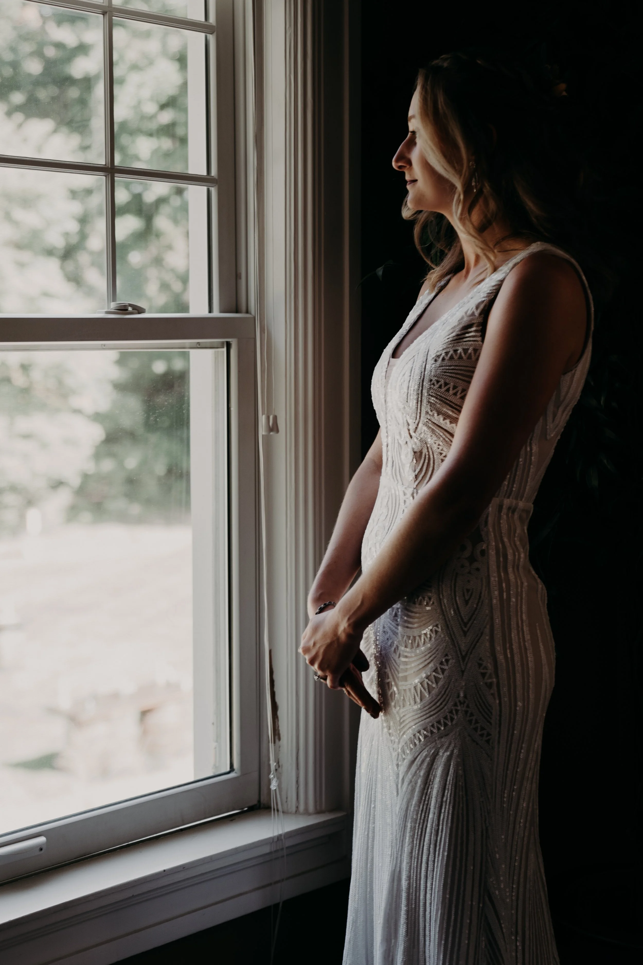 Woman in a white detailed dress standing inside by a window, looking outside with a serene expression.