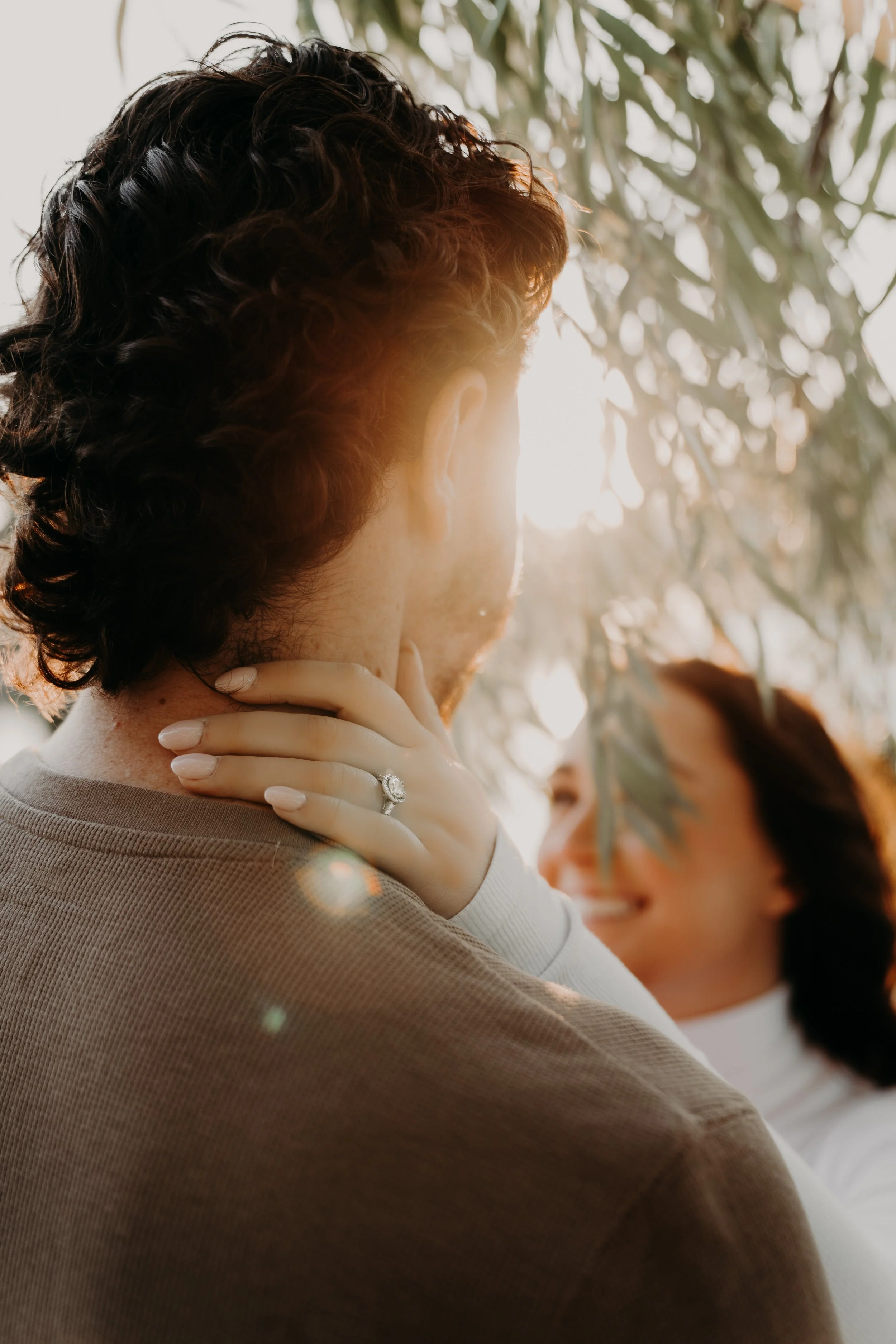 Close-up of a woman with curly dark hair, wearing a ring on her finger, touching her neck, with sunlight and leaves in the background.