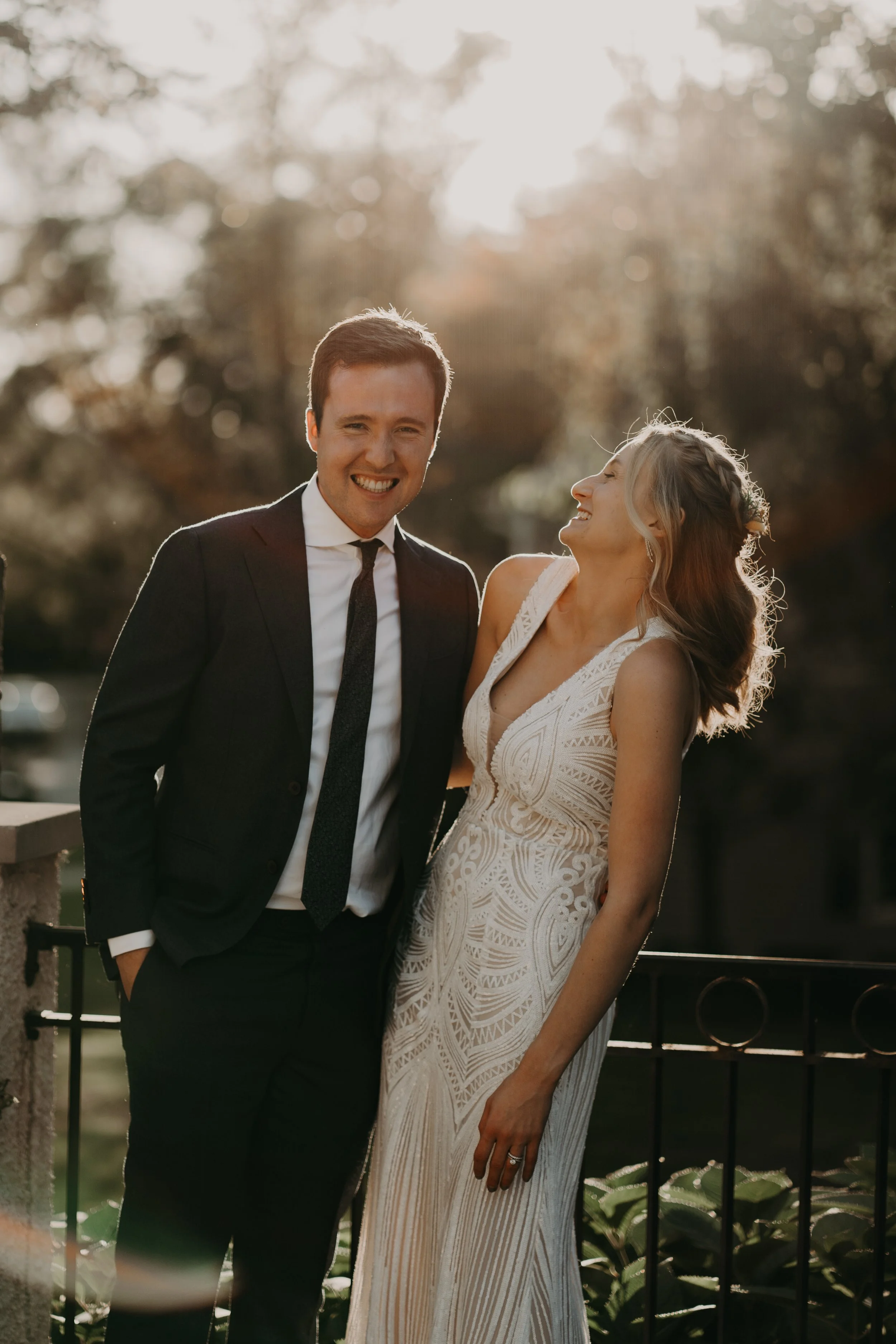 A smiling man in a black suit and tie stands next to a woman in a white, sleeveless, patterned dress, both on a balcony with a black fence. The woman is laughing and looking at the man. The sunlight is shining through trees in the background.
