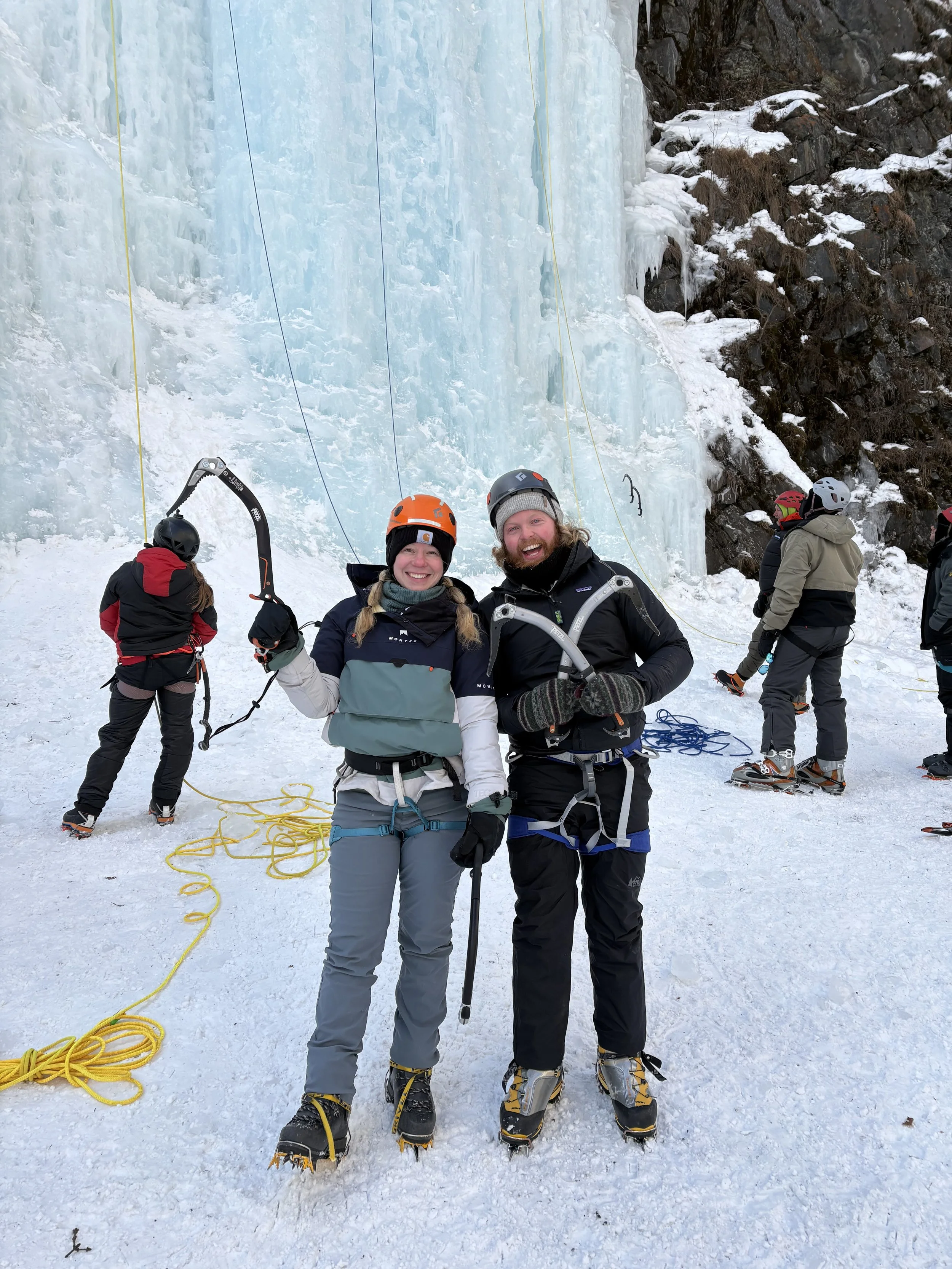 People ice climbing in a snowy mountain area, wearing helmets and winter gear, with one person smiling and holding climbing equipment, others preparing or resting nearby.