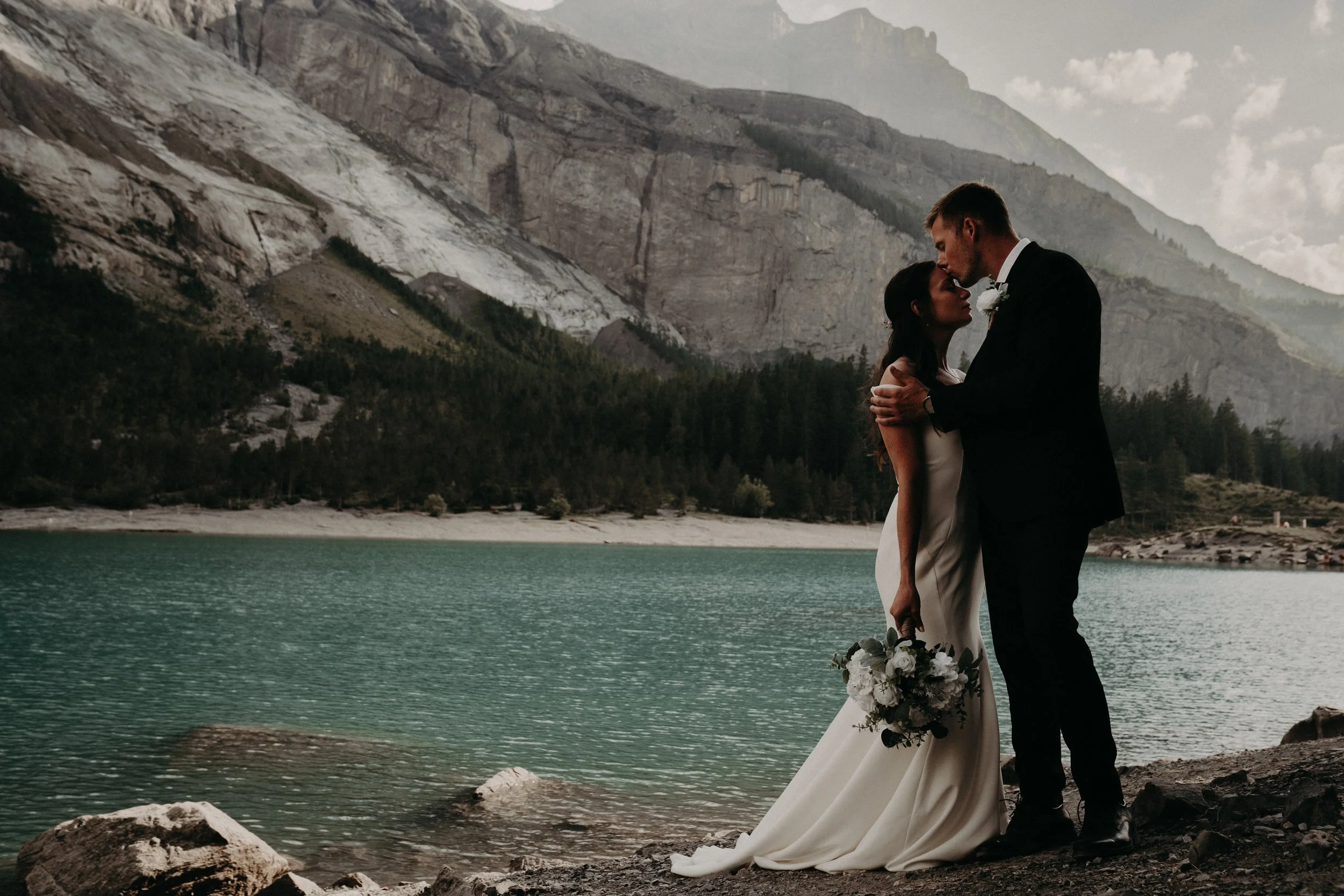 A bride and groom embracing by a lake, with mountains and a cloudy sky in the background.