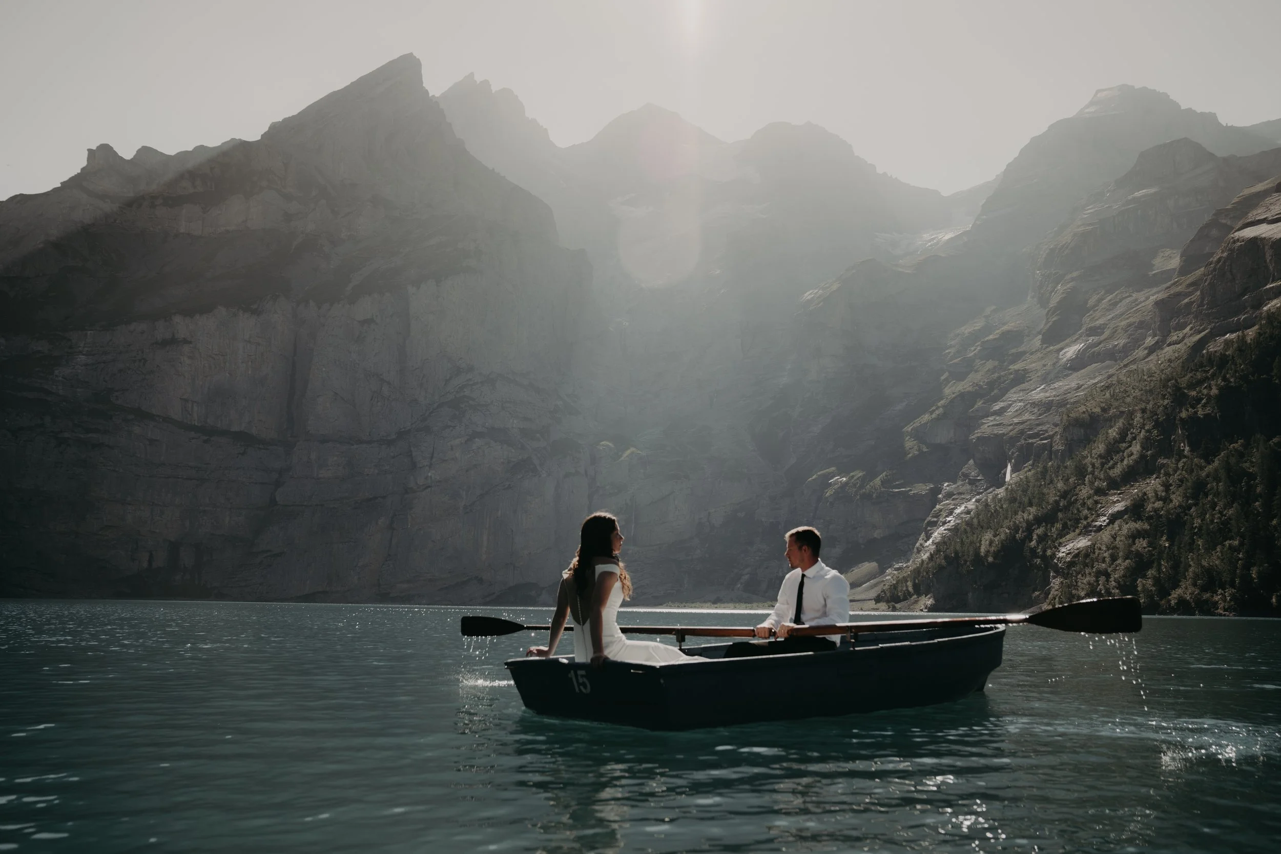 A couple in formal dress is in a boat on a lake with mountains in the background, illuminated by sunlight.