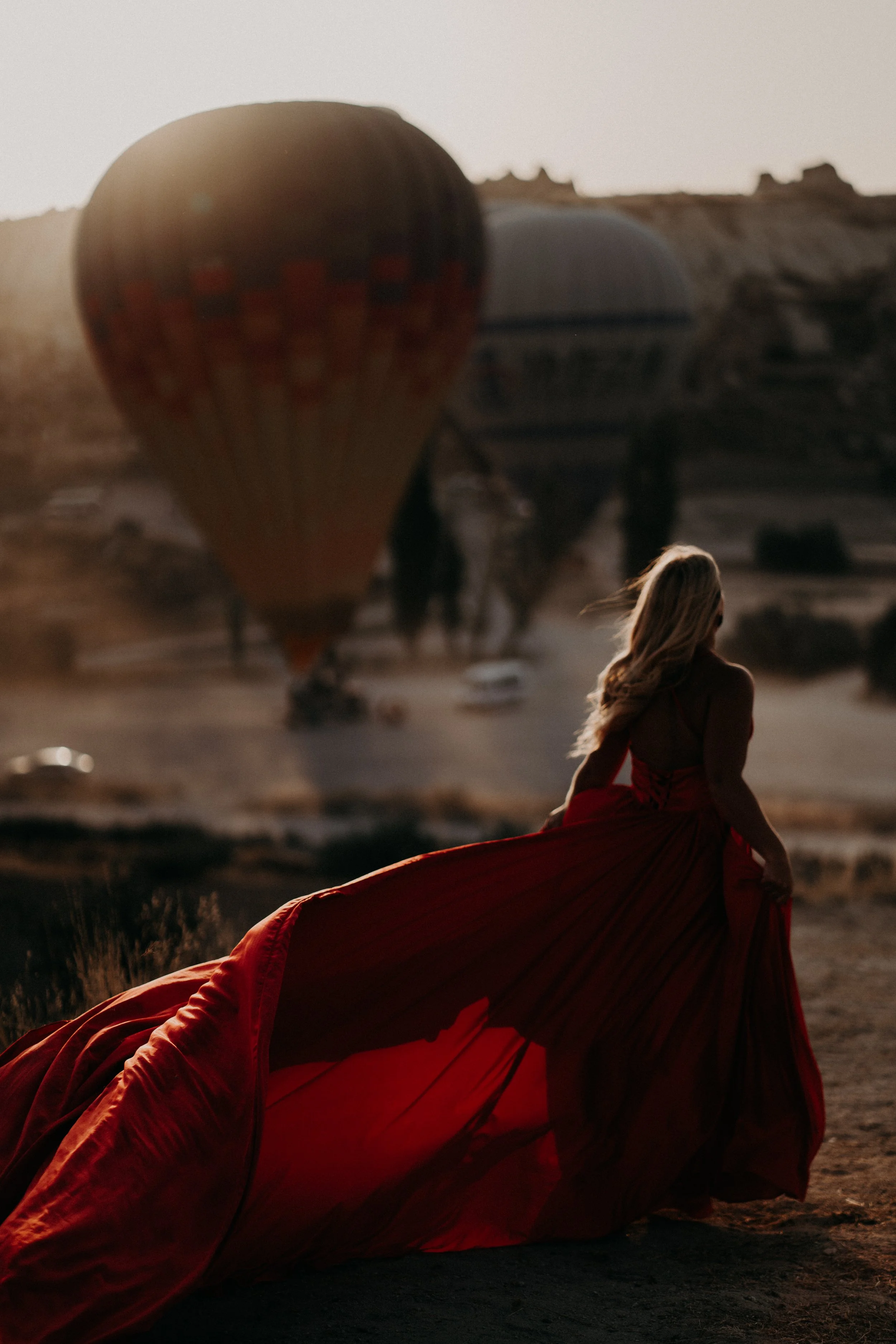 A woman in a red dress sitting outdoors near a hot air balloon during sunset.