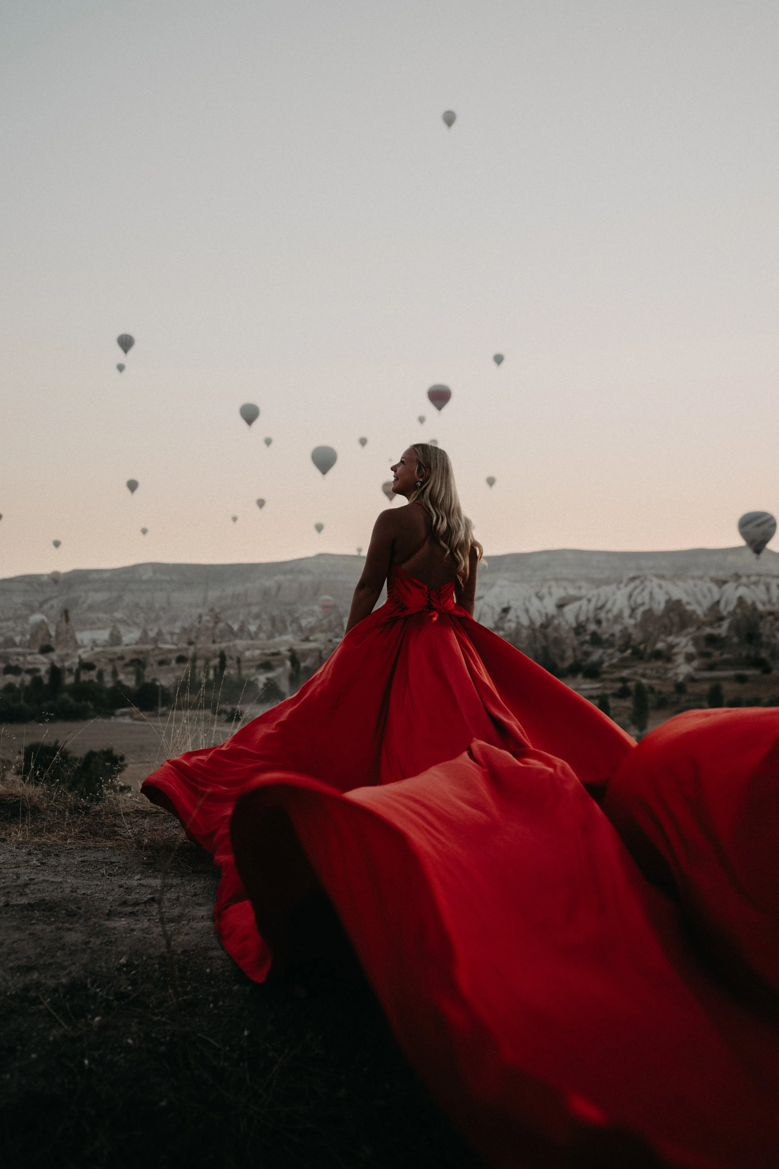 A woman in a red dress standing outdoors during sunset, with hot air balloons flying in the sky in the background.