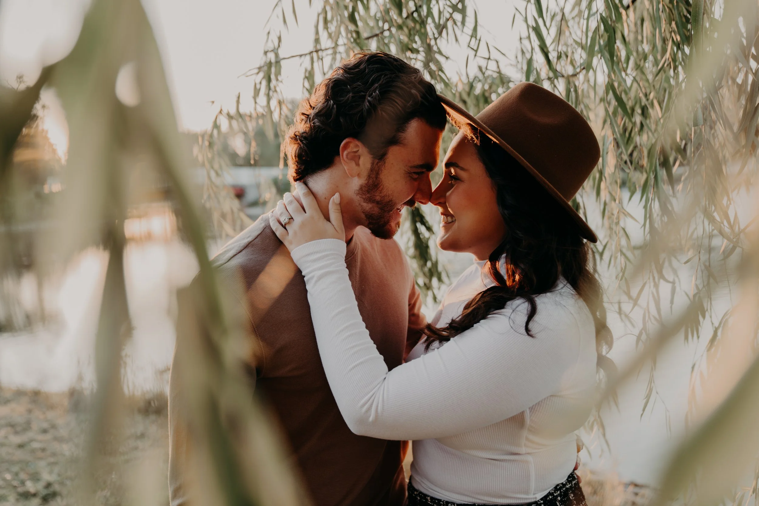 A couple is close together, touching foreheads, smiling and embracing amid tall leafy plants during sunset.