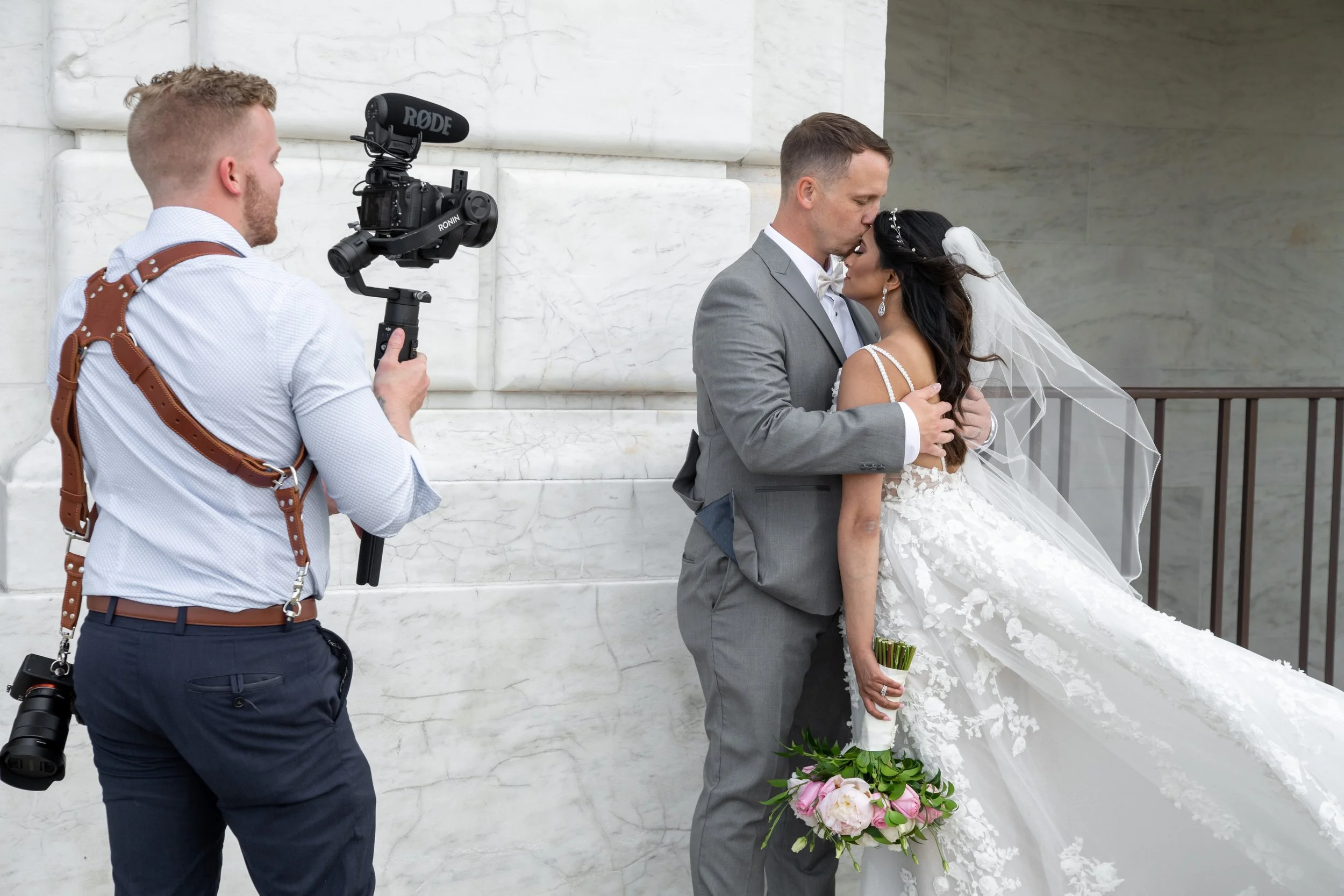 A bride and groom share a kiss during their wedding ceremony while a photographer captures the moment. The bride holds a bouquet of pink and white flowers, and the groom is dressed in a gray suit with a white bow tie. A camera operator with a shoulder strap and a camera stabilizer films the scene against a white marble wall.