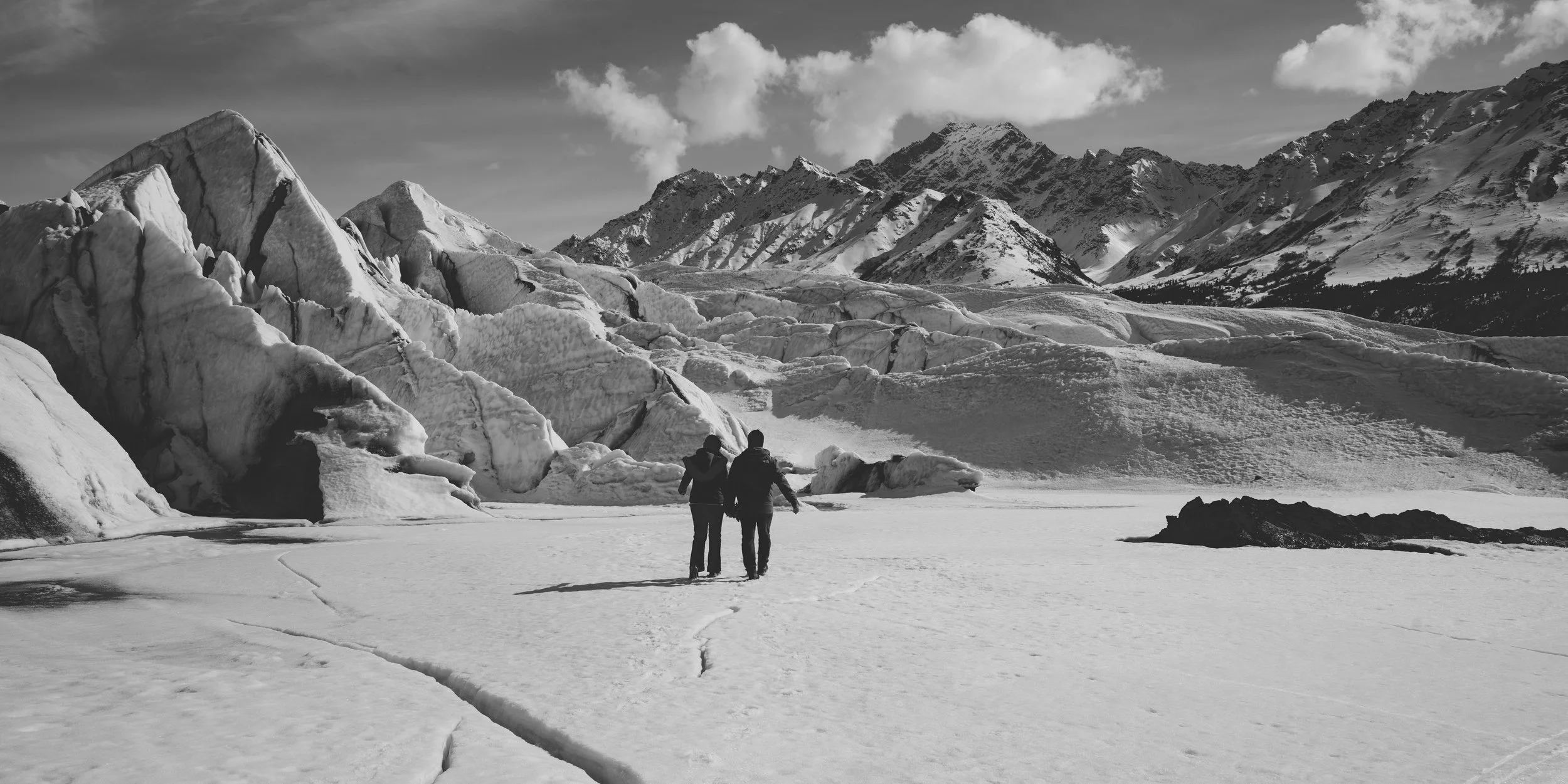 Two people holding hands walking on snow-covered terrain with large ice formations and mountain peaks in the background, in black and white.