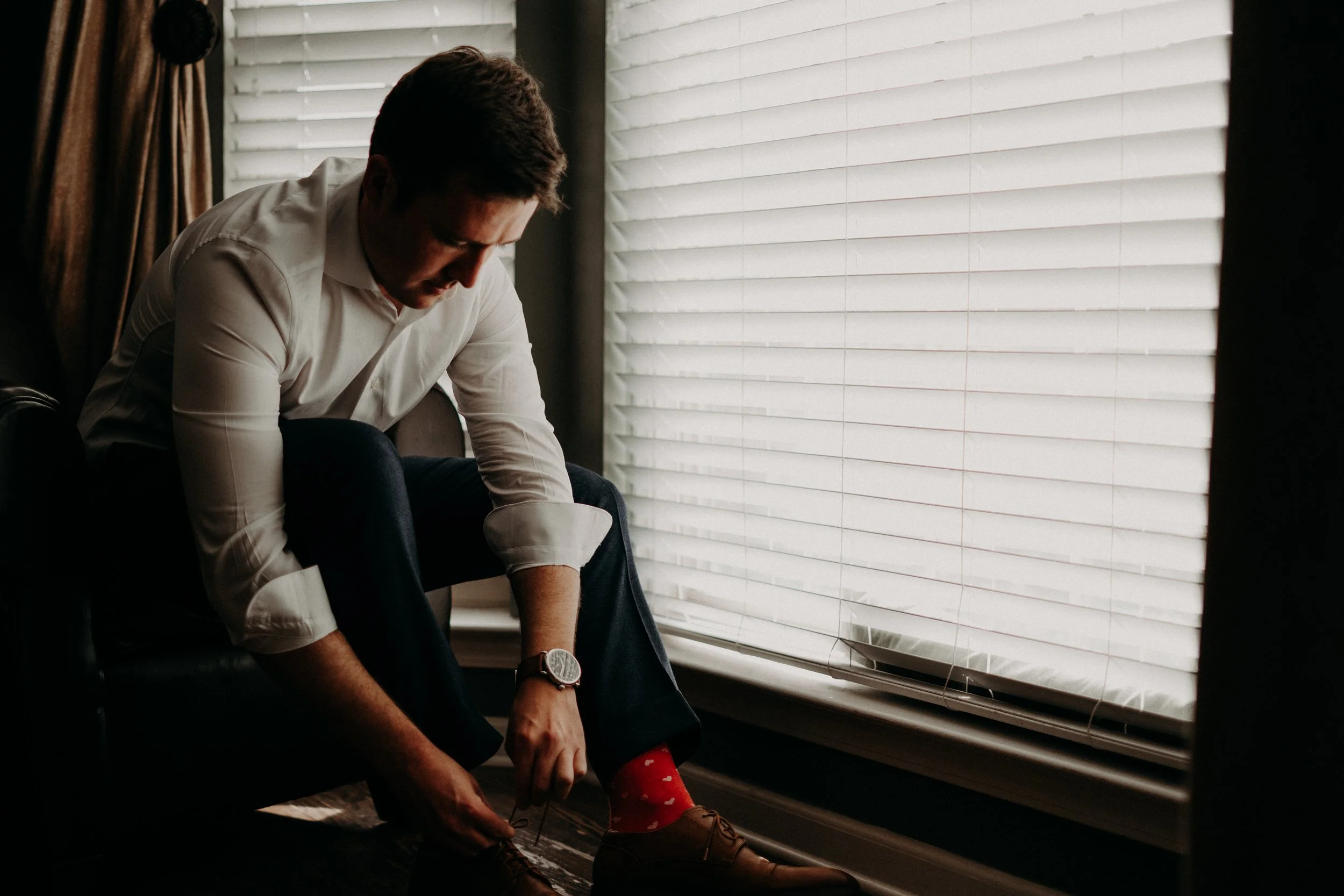 A man in a white shirt and dark pants sits by a window with white blinds, leaning forward and tying the shozen of a young girl wearing red socks with heart patterns. The man is focused on his task, and the scene is softly lit by natural light coming through the blinds.