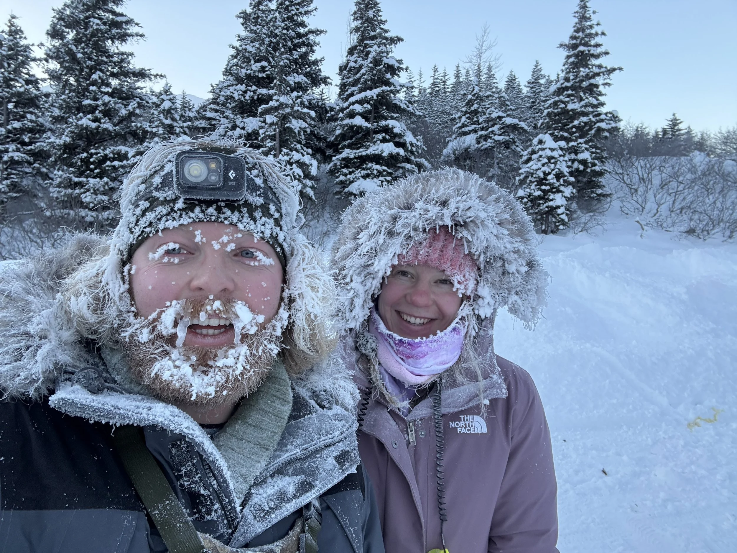 A close-up of two smiling women in winter clothing, partly covered in snow, with snow-covered trees and a clear blue sky in the background.