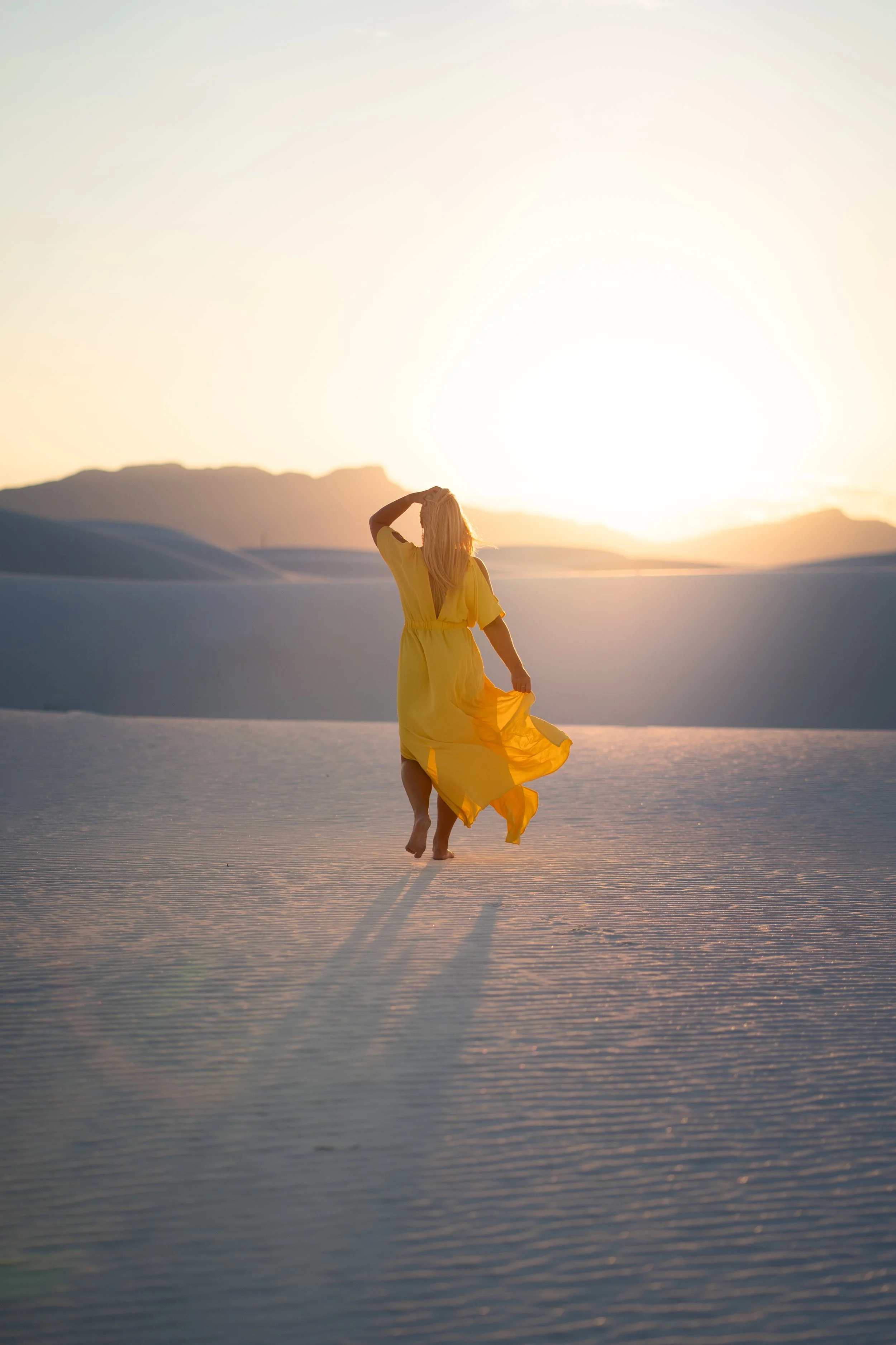 A woman in a yellow dress walking barefoot on a flat, sandy landscape during sunset, with mountain silhouettes in the background.