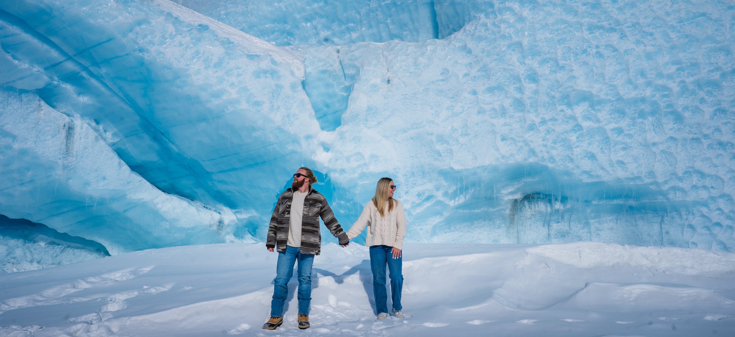 A man and woman holding hands standing on snow in front of a large blue ice wall or glacier.  Elopement photography