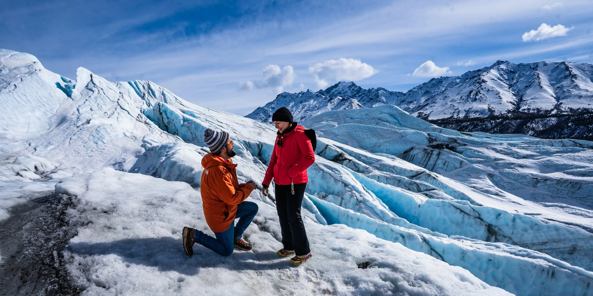 Two people, one kneeling and proposing, on snowy terrain with glaciers and snow-covered mountains in the background.