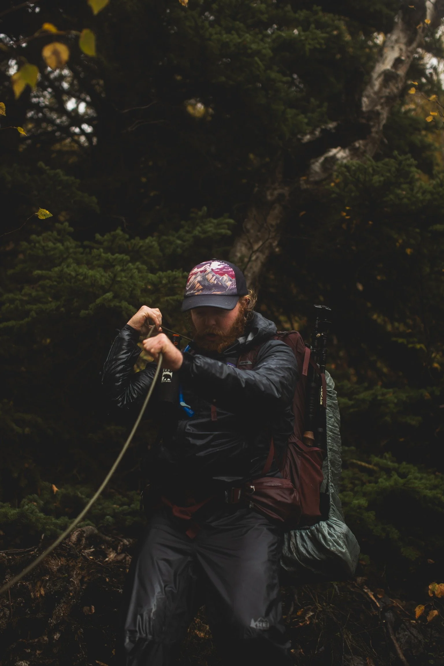 A man with a beard, wearing a camouflage baseball cap and outdoor gear, is adjusting a rope or strap in a forest setting with dense trees and foliage.