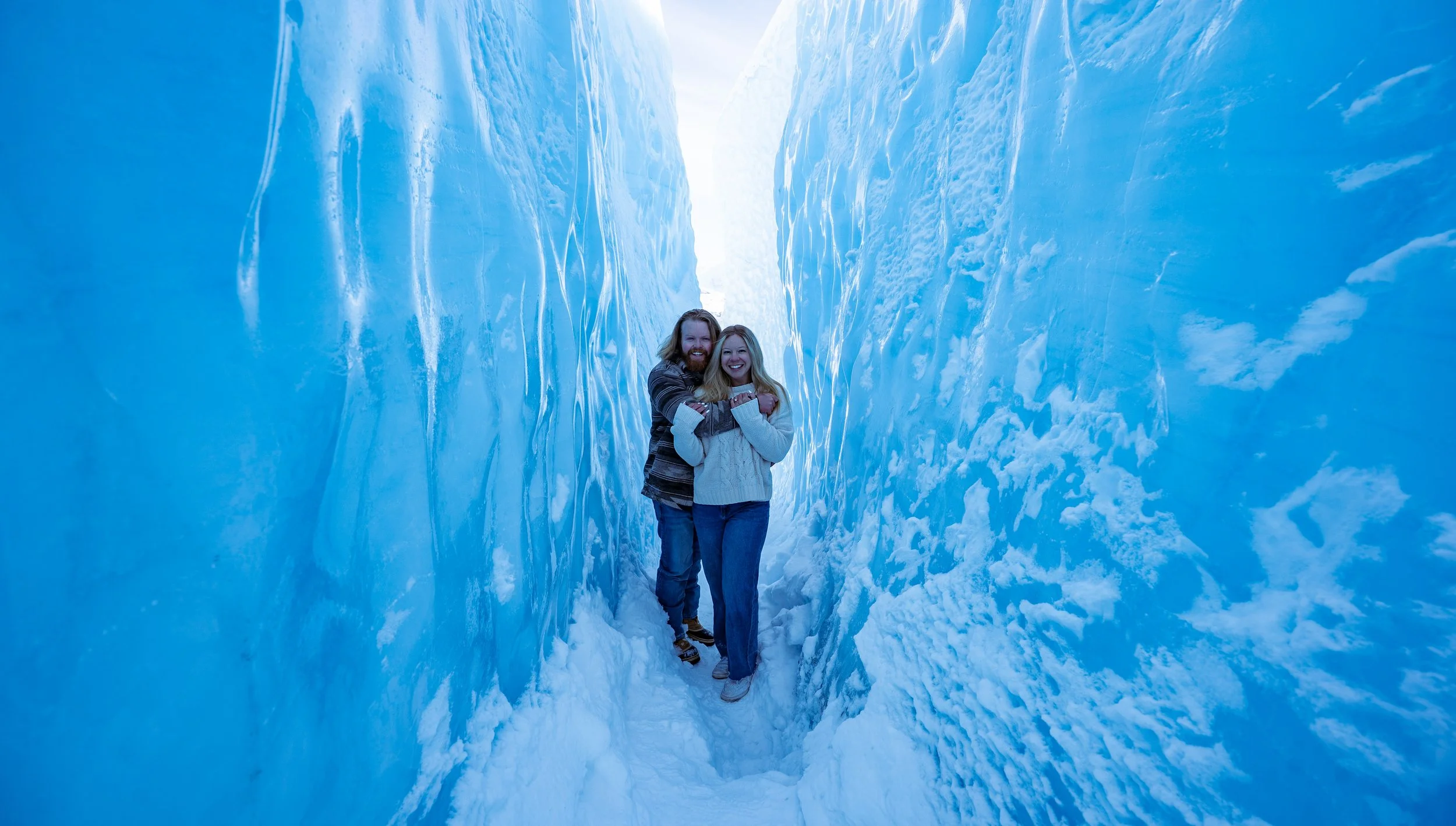 Two smiling people, a man with a beard and a woman, standing inside a narrow, icy crevasse between tall ice walls, holding each other.