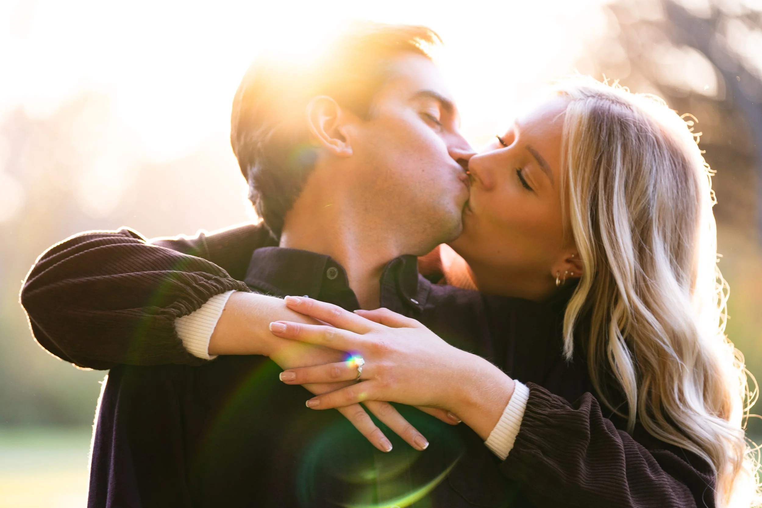 A young man and woman sharing a kiss outdoors in sunlight, with the woman’s arms around the man's neck.