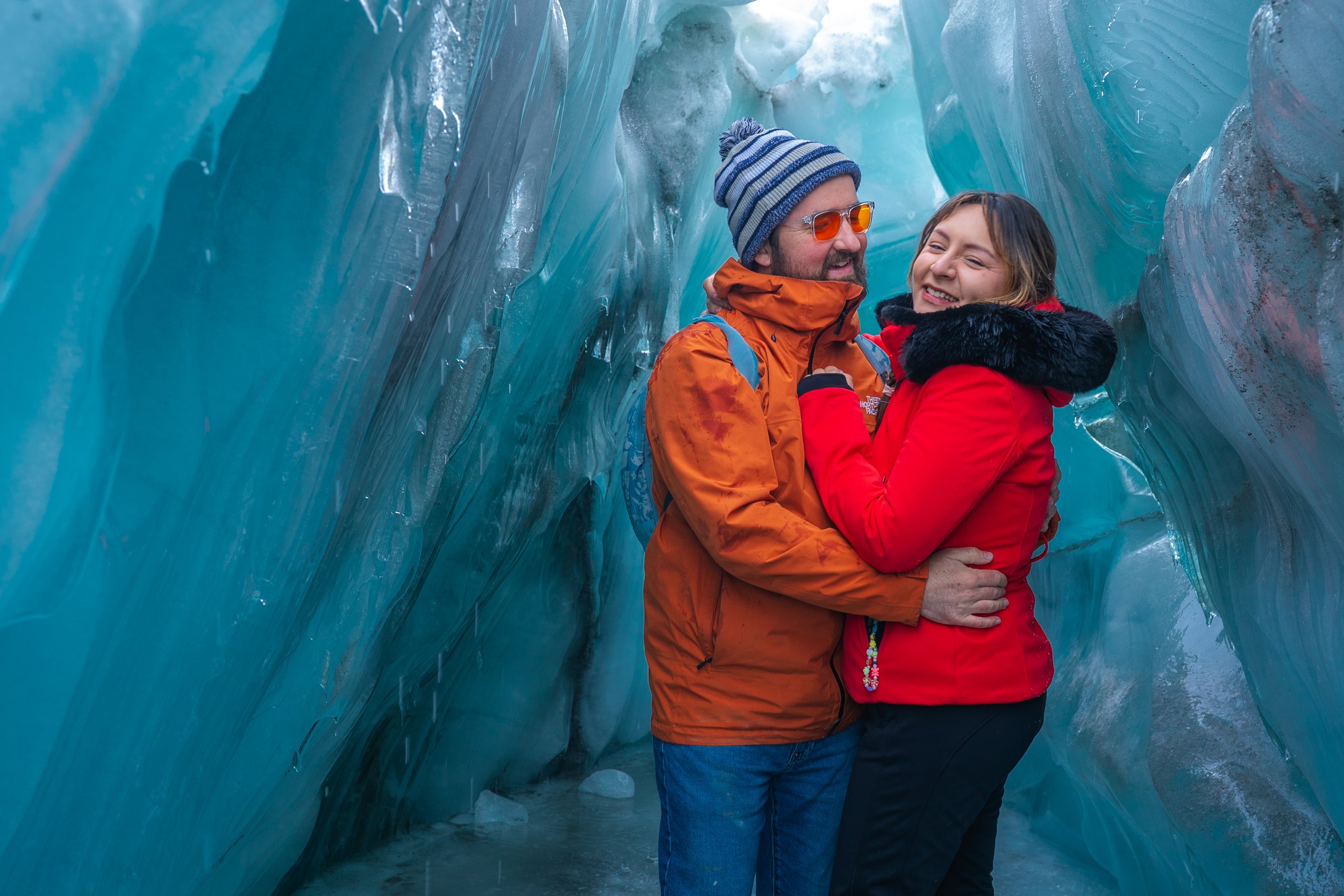 A couple hugging inside a blue ice cave, dressed in orange and red winter jackets.