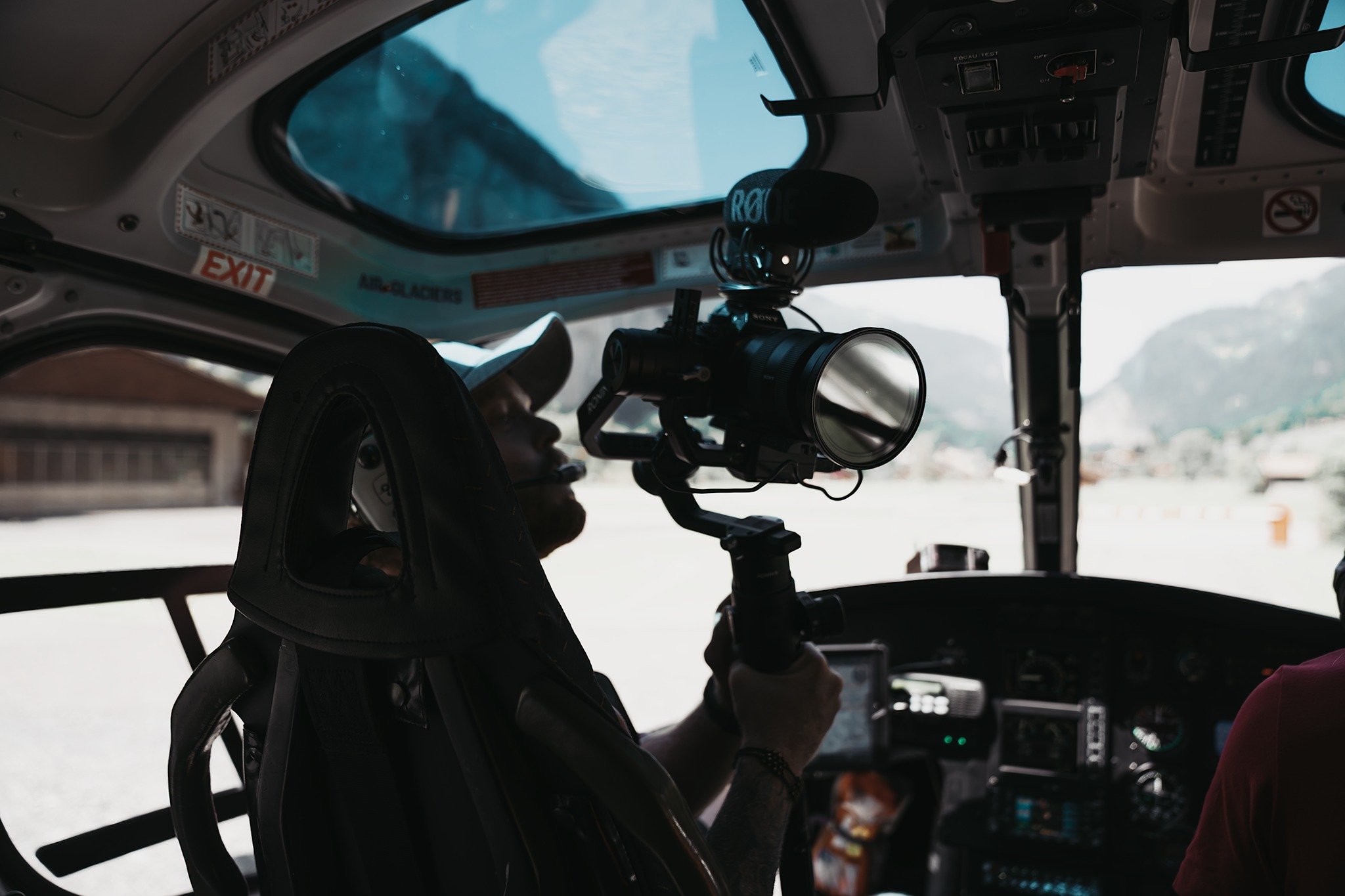 The image shows the interior of a vehicle, possibly an emergency or patrol vehicle, with a camera or scope mounted near the rearview mirror. The view through the windshield reveals a mountainous landscape under a partly cloudy sky.