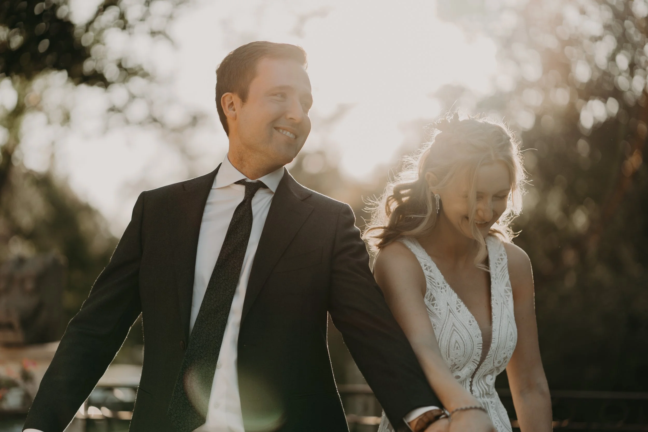 A smiling couple, a man in a suit and a woman in a white dress, holding hands outdoors in sunlight with trees in the background.