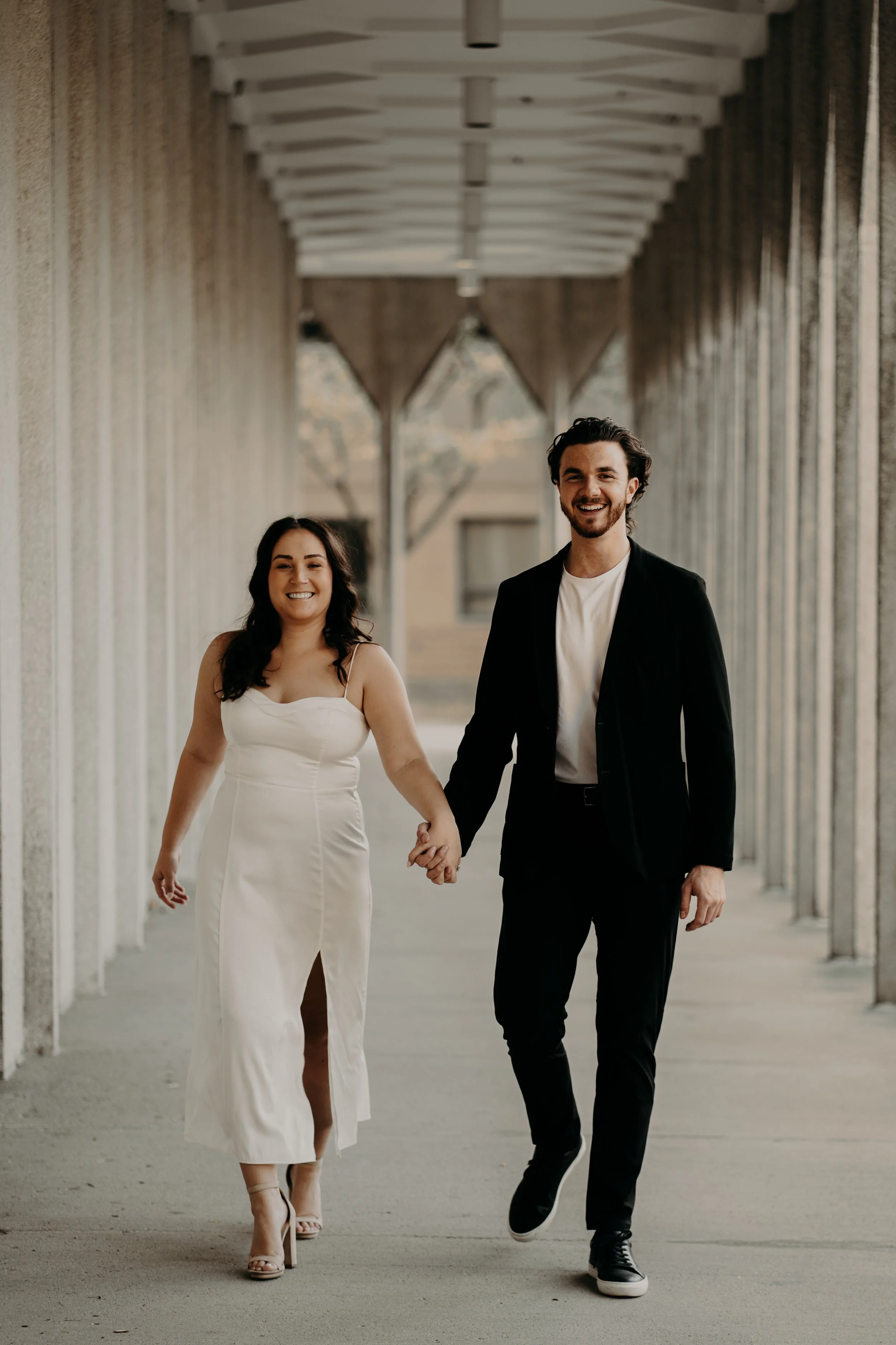 A smiling couple walking hand in hand in an outdoor corridor with columns and beams overhead.