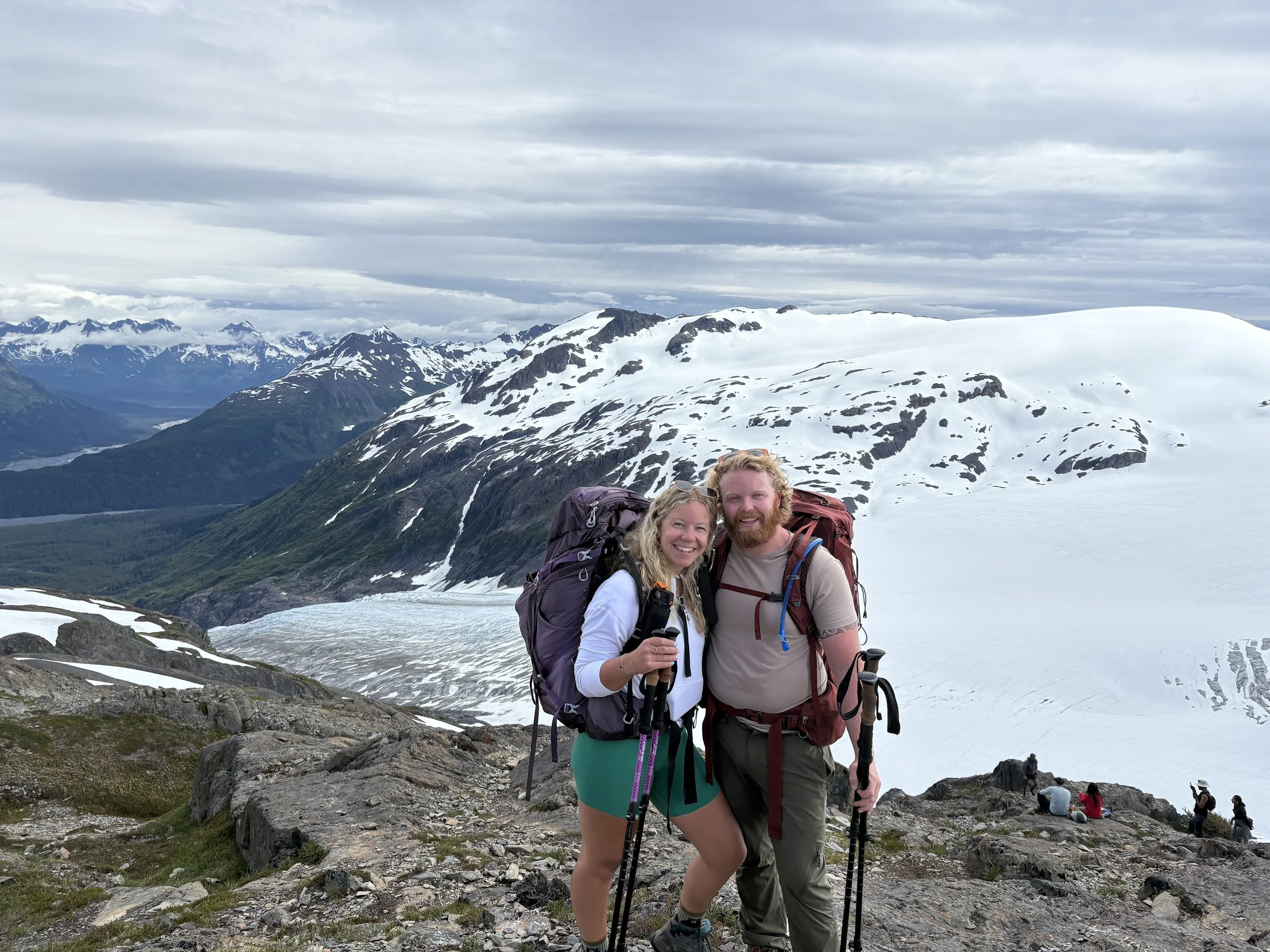 A smiling couple dressed in hiking gear standing on a rocky area on a snow-capped mountain, with a scenic view of mountain peaks and clouds in the background.