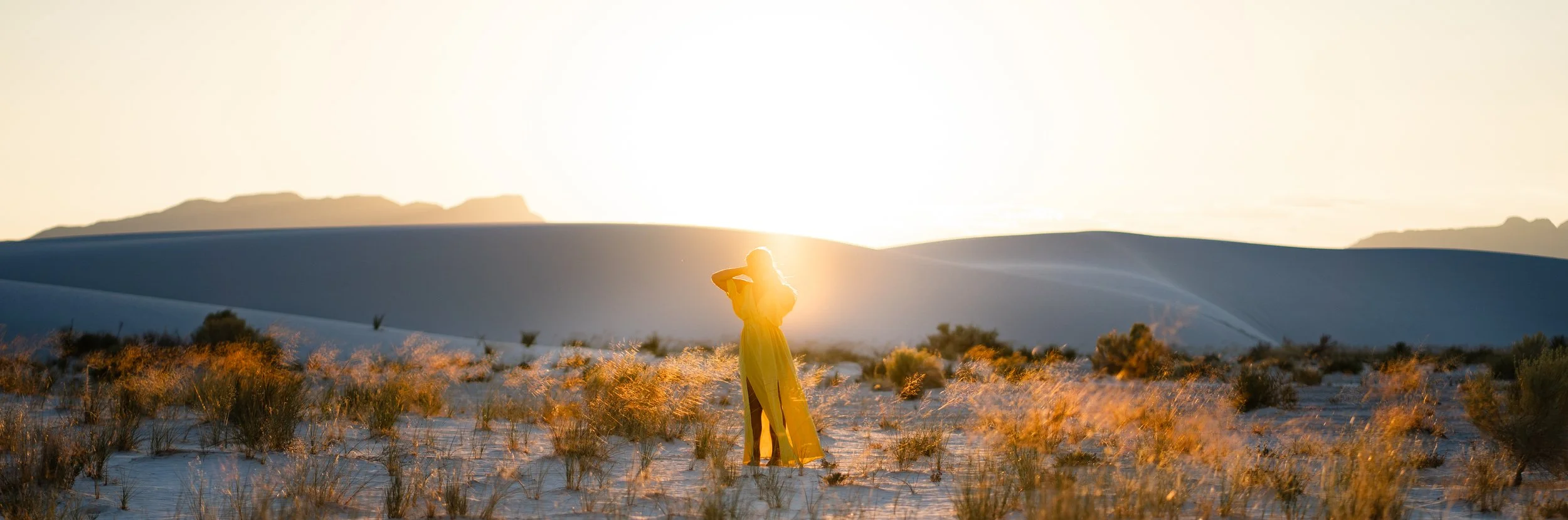 A person in a flowing yellow dress standing in a desert landscape at sunset, with the sun behind them and hills in the background.
