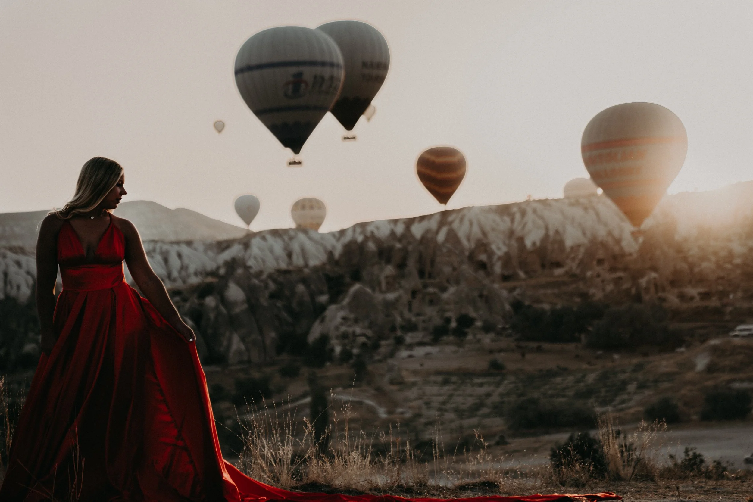 A woman in a flowing red dress standing outdoors at sunset, with several hot air balloons floating in the sky behind her over rocky terrain.