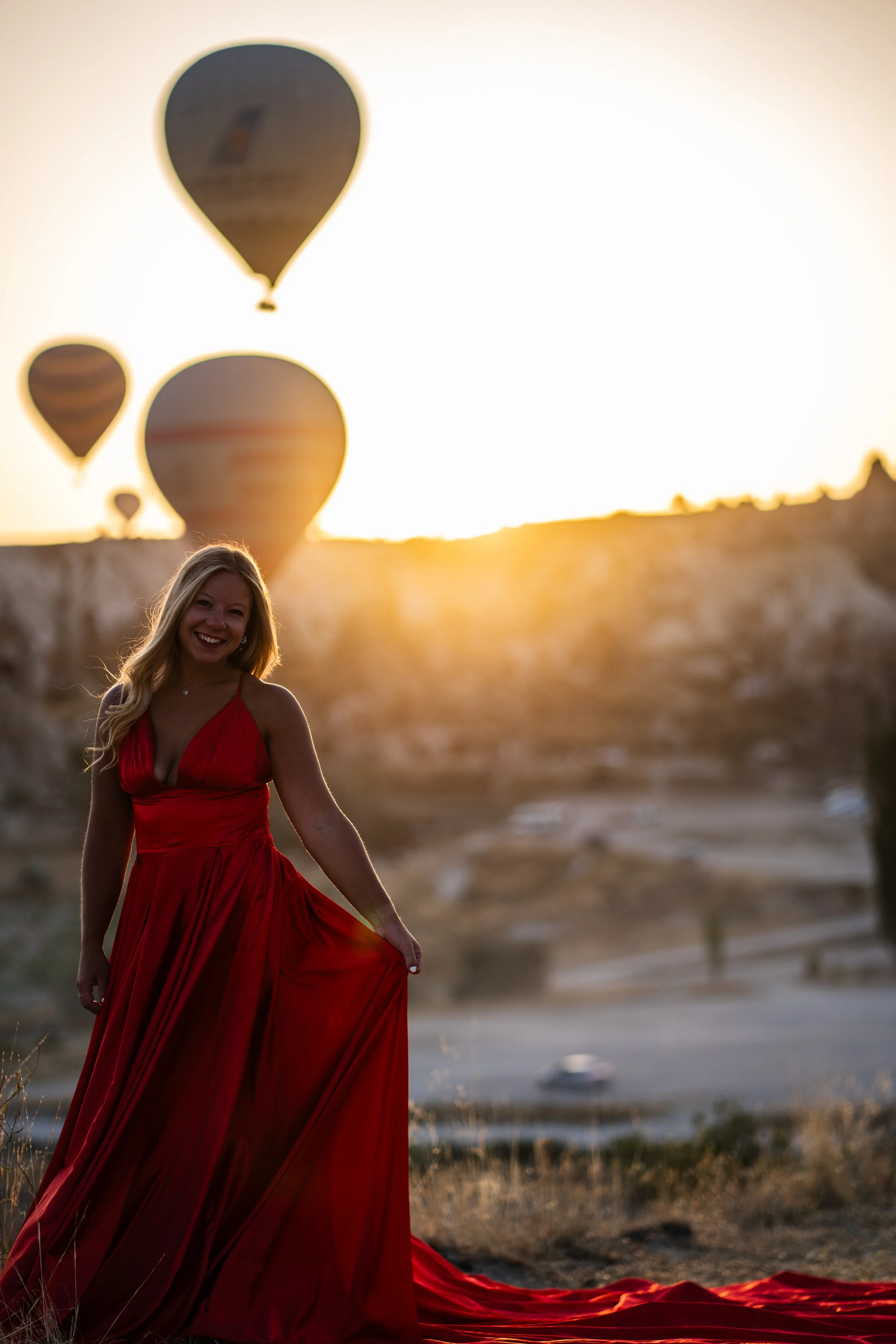A woman in a long red dress smiling outdoors during sunset with hot air balloons in the background.