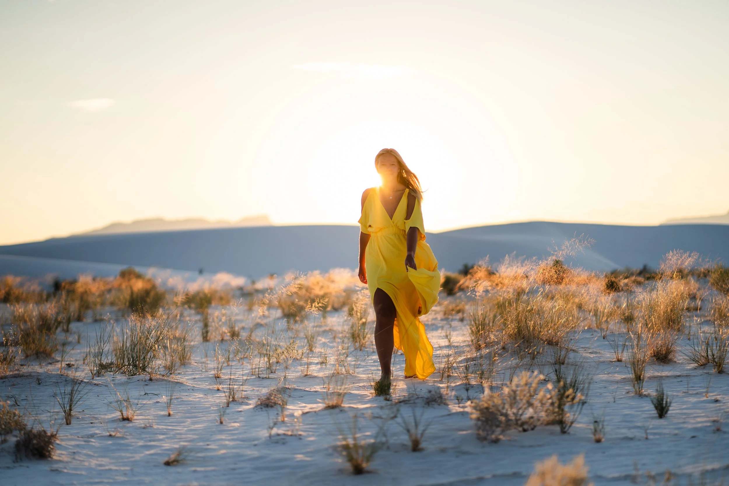 A woman in a yellow dress walking through a sandy field with sparse vegetation at sunset, with mountains in the background.